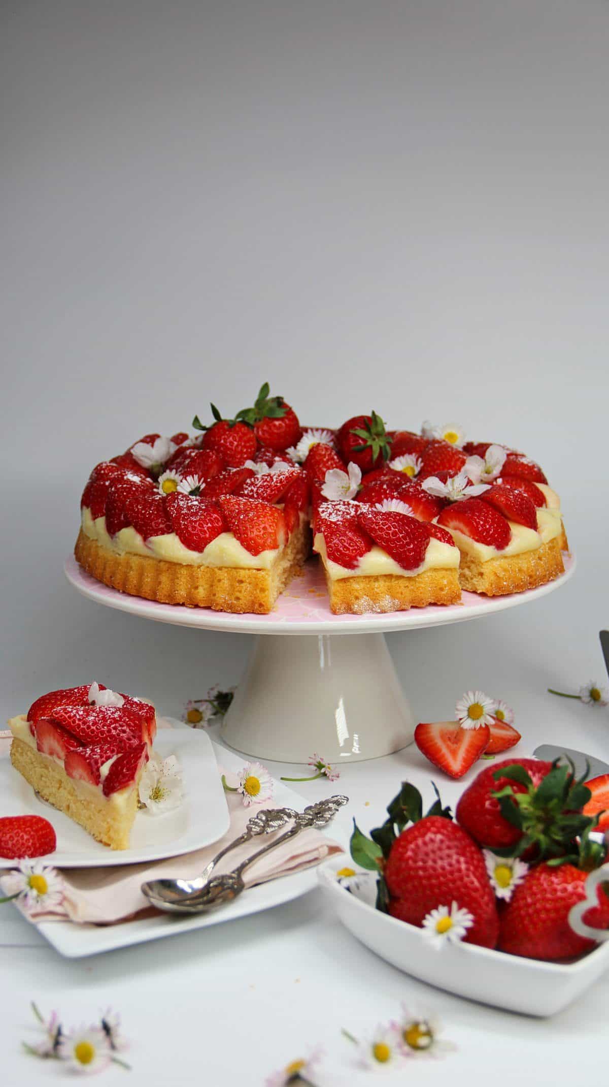 Erdbeerkuchen auf einer weißen Tortenplatte mit vielen frischen Erdbeeren und Blüten dekoriert