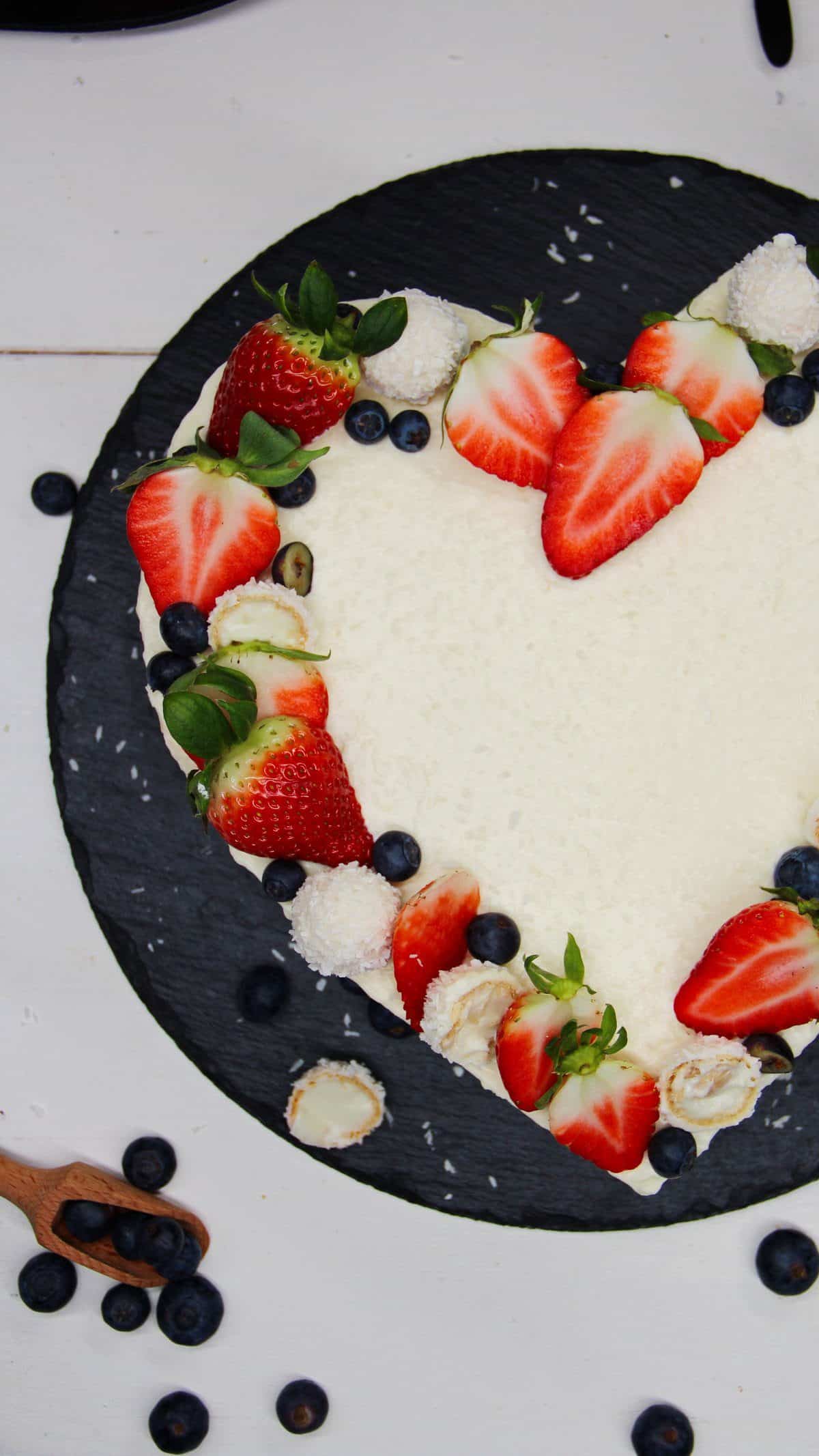 Raffaello Torte in Herzform auf einer schwarzen Schieferplatte. Mit Raffaellos, Erdbeeren und Blaubeeren dekoriert. Nahaufnahme.