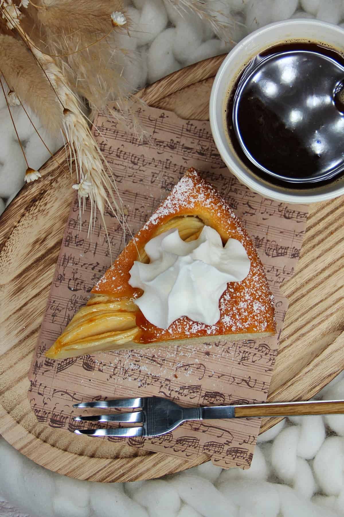 Versunkener Apfelkuchen, Kuchenstück mit Sahne und einer Tasse Kaffee auf einem kleinen Holz-Servierbrett