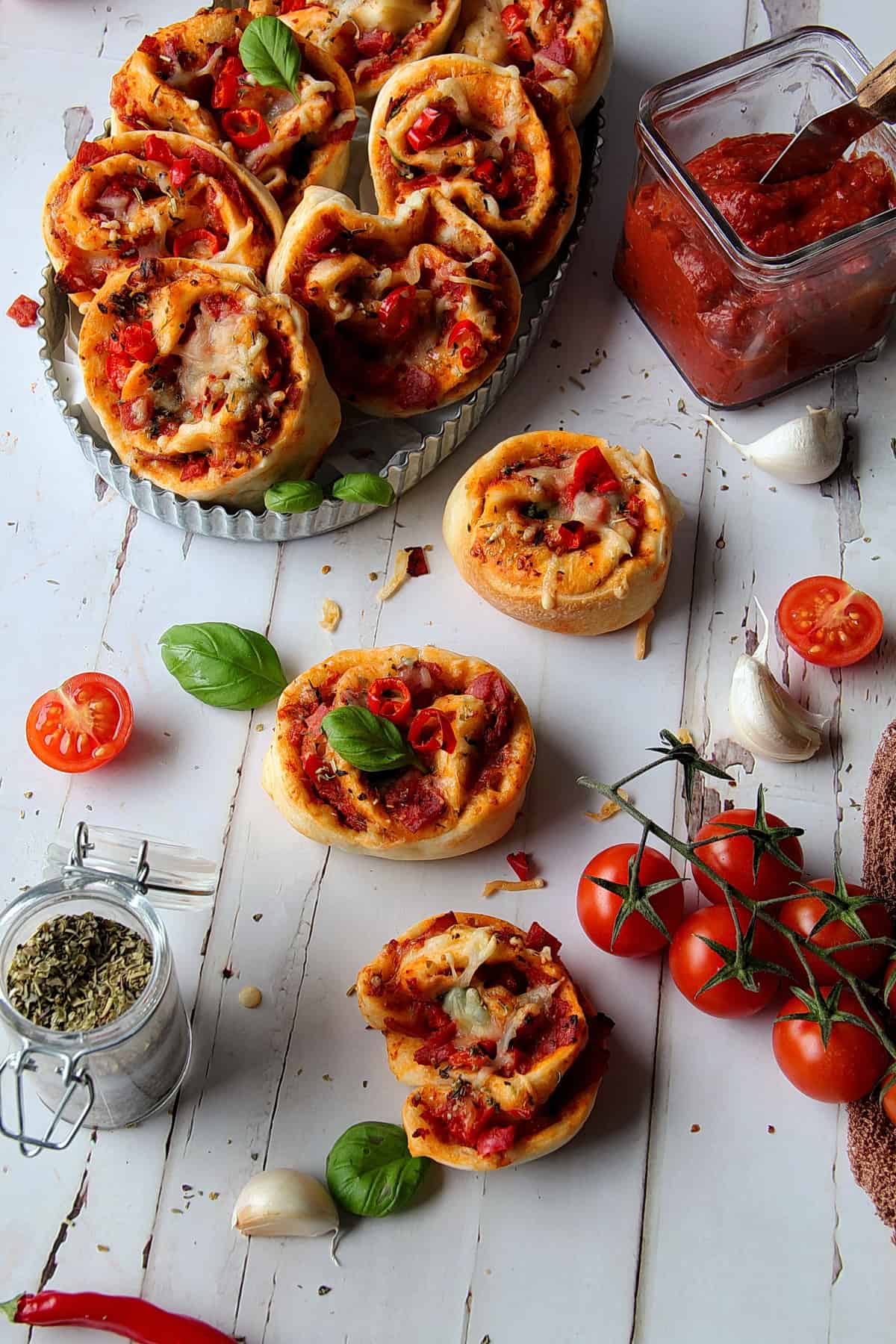 Pizzaschnecken in einer Schale und auf dem Tisch verteilt, neben Tomaten, Knoblauch und Basilikum