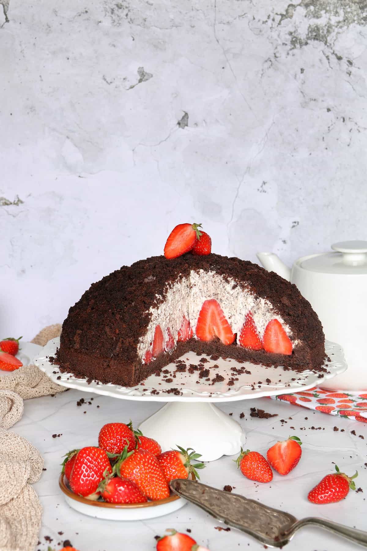 Maulwurfkuchen mit Erdbeeren auf einem weißen Tortenständer, im Anschnitt. Im Vordergrund ein Schälchen frischer Erdbeeren und ein antiker Tortenheber.