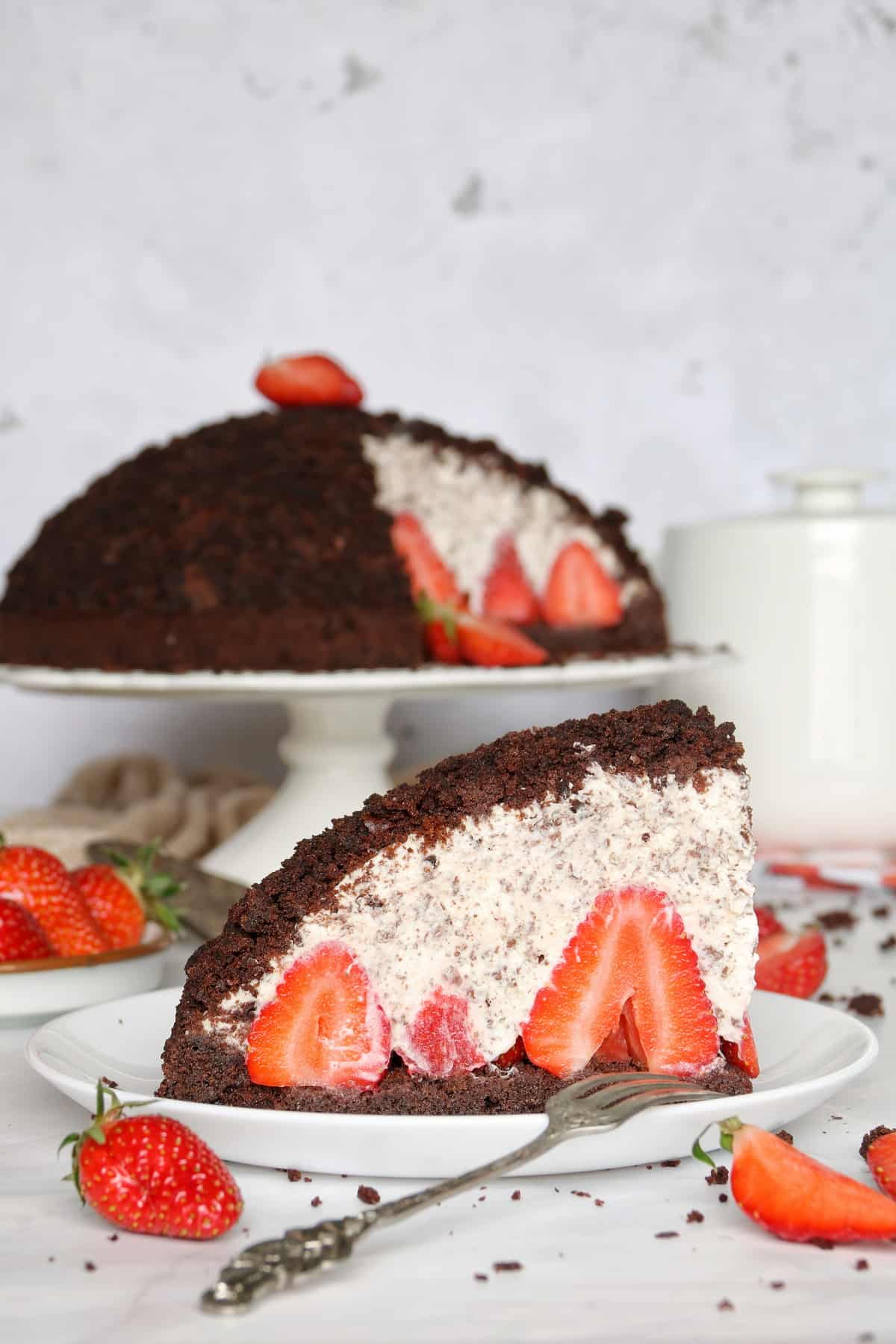 Maulwurfkuchen mit Erdbeeren auf einem weißen Tortenständer, im Anschnitt. Kuchen ist auf einem Tortenständer unscharf im Hintergrund zu sehen.
