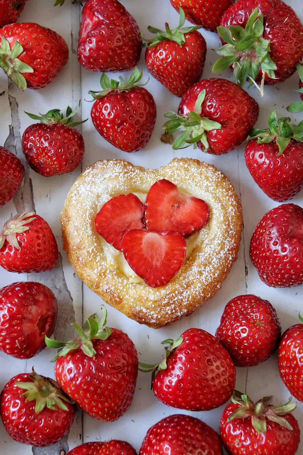 Blätterteig-Herzen mit Erdbeeren auf einem weißen, abgeblätterten Tisch umringt von frischen Erdbeeren