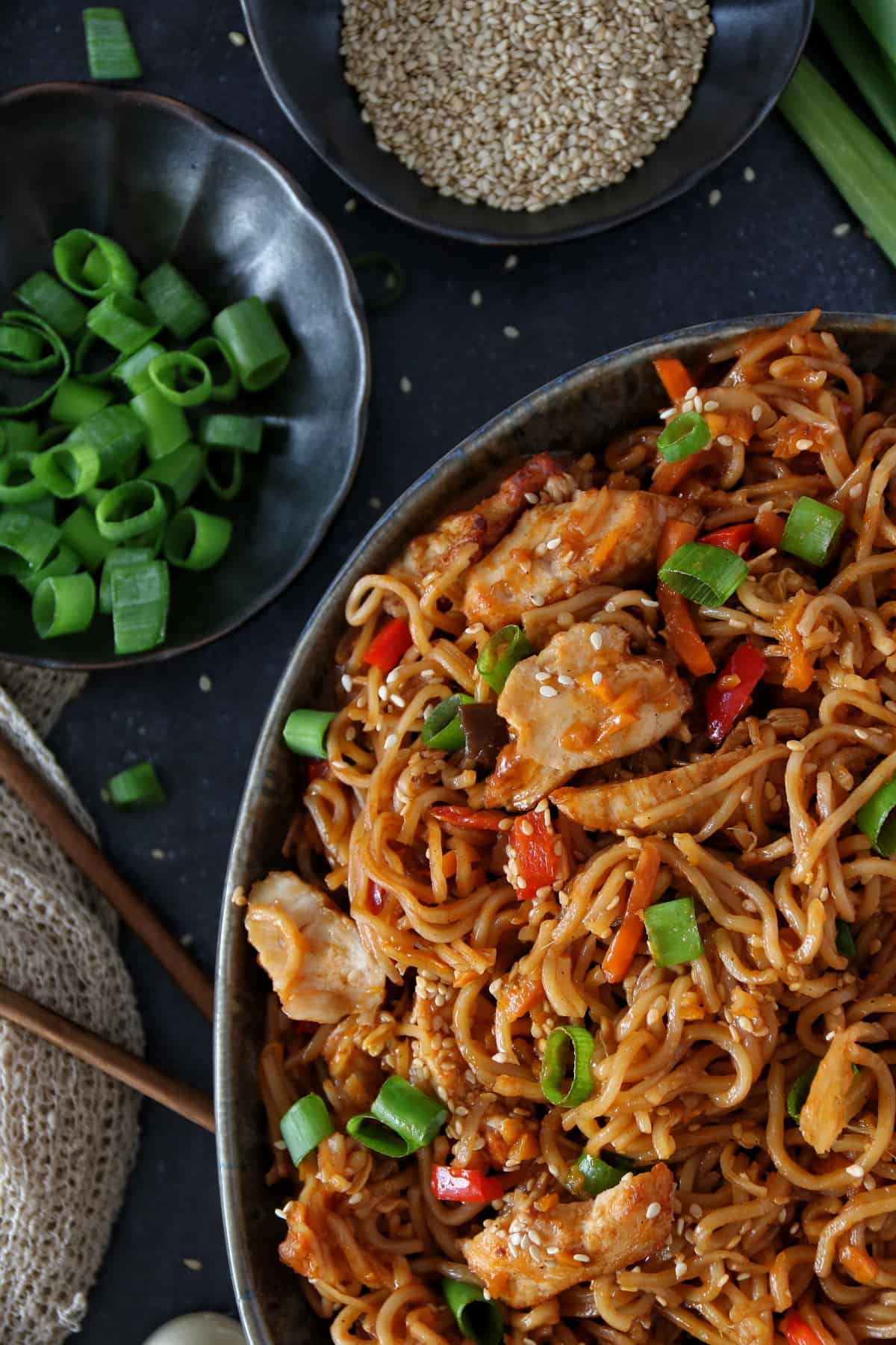 All-in-One-Gericht: Chinesische Nudeln mit Gemüse und Hähnchen aus dem Thermomix® in einer schwarzen Schüssel. Daneben zwei Schälchen mit Schnittlauch und Sesamsamen.