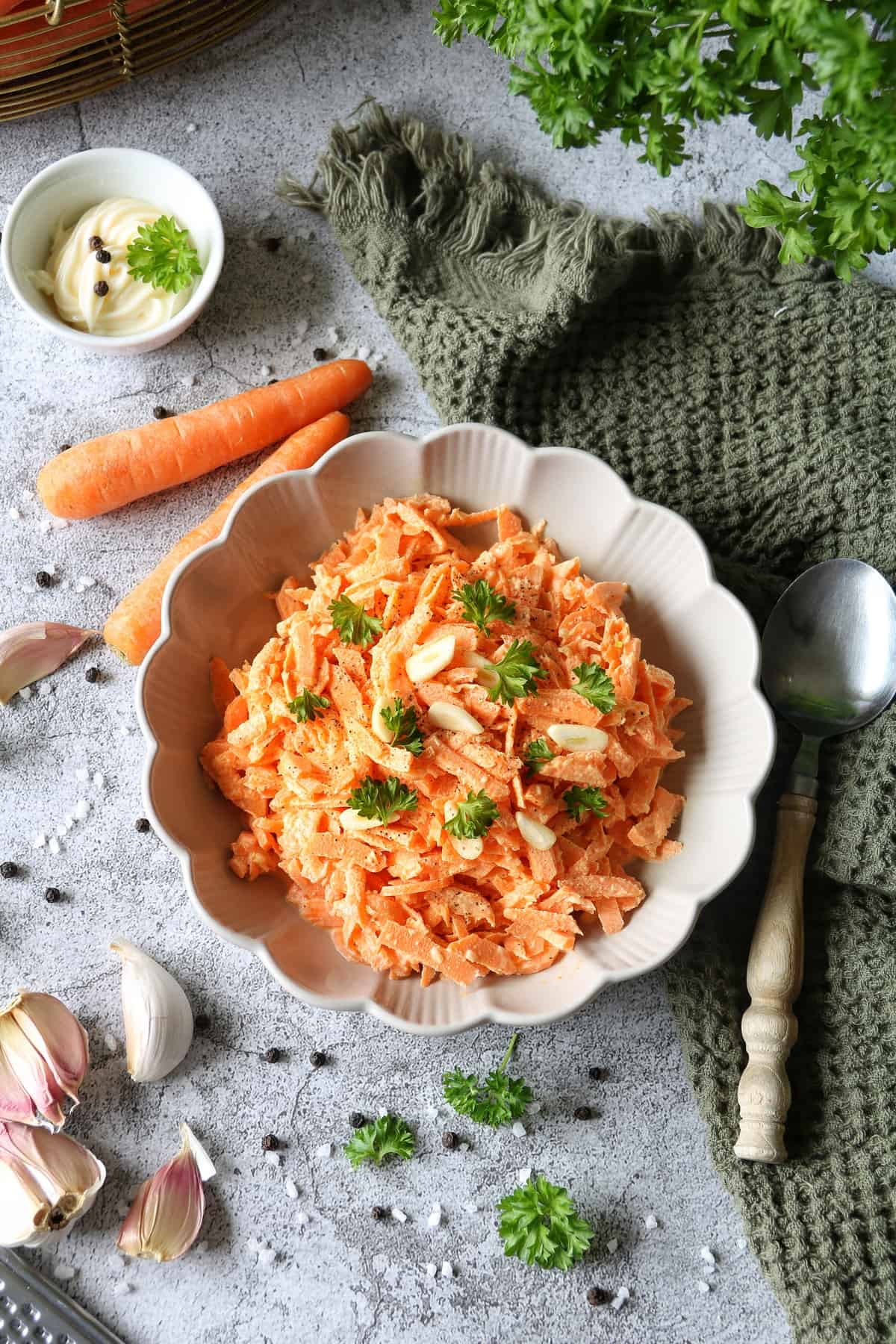 Einfacher Karottensalat mit Schmand und Mayonnaise in einem Teller mit Wellenrand. Daneben liegt ein grünes Waffelhandtuch und ein Löffel.