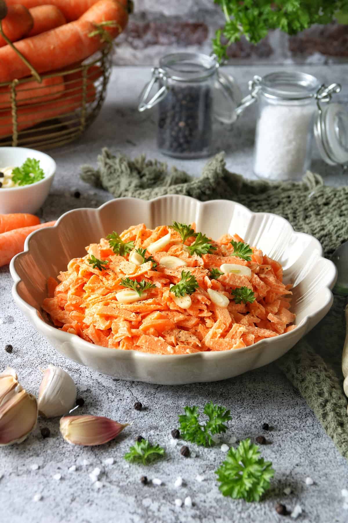 Einfacher Karottensalat mit Schmand und Mayonnaise in einem Teller mit Wellenrand. Daneben liegt ein grünes Waffelhandtuch und ein Löffel. Im Hintergrund sind Möhren in einem Drahtkorb.
