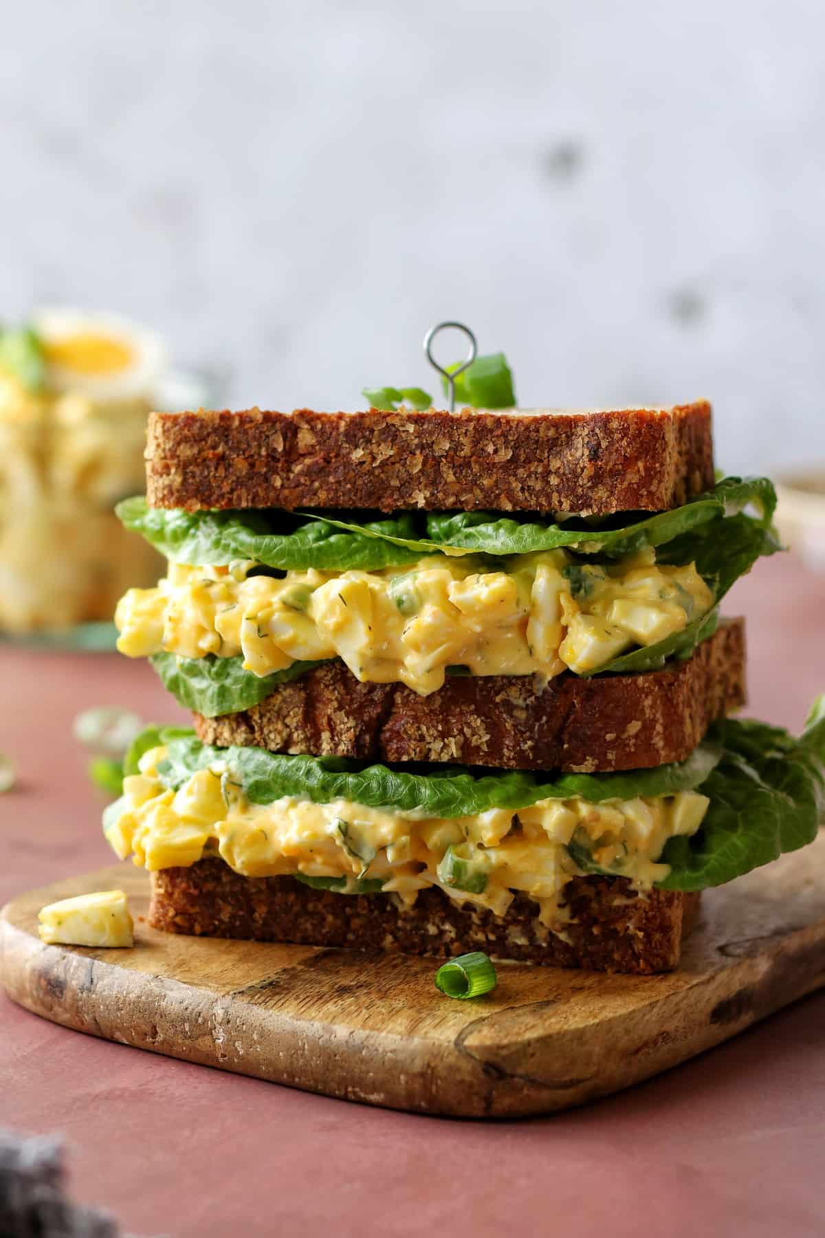 Der Eiersalat ist als Füllung für einen Sandwich-Burger zu sehen. 3 dicke Brotscheiben sind mit Eiersalat und Blattsalat gestapelt und werden mit einem Spieß zusammengehalten.