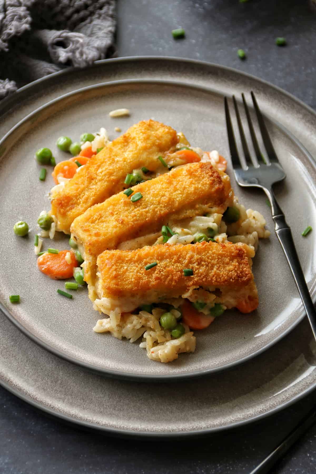 Tellerbild vom Fischstäbchen-Auflauf. Reis und Gemüse liegen mit 3 Fischstäbchen auf einem grauen Teller. Daneben liegt eine schwarze Gabel.