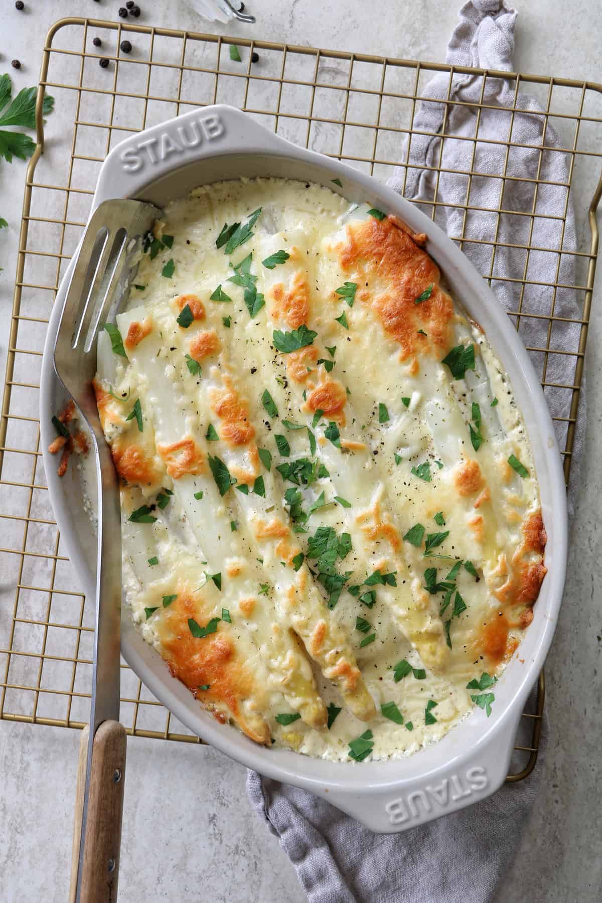 Ein köstlicher Spargelauflauf ist zu sehen, mit weißem Spargel in einer grauen STAUB Auflaufform, die auf einem goldenen Abkühlgitter steht