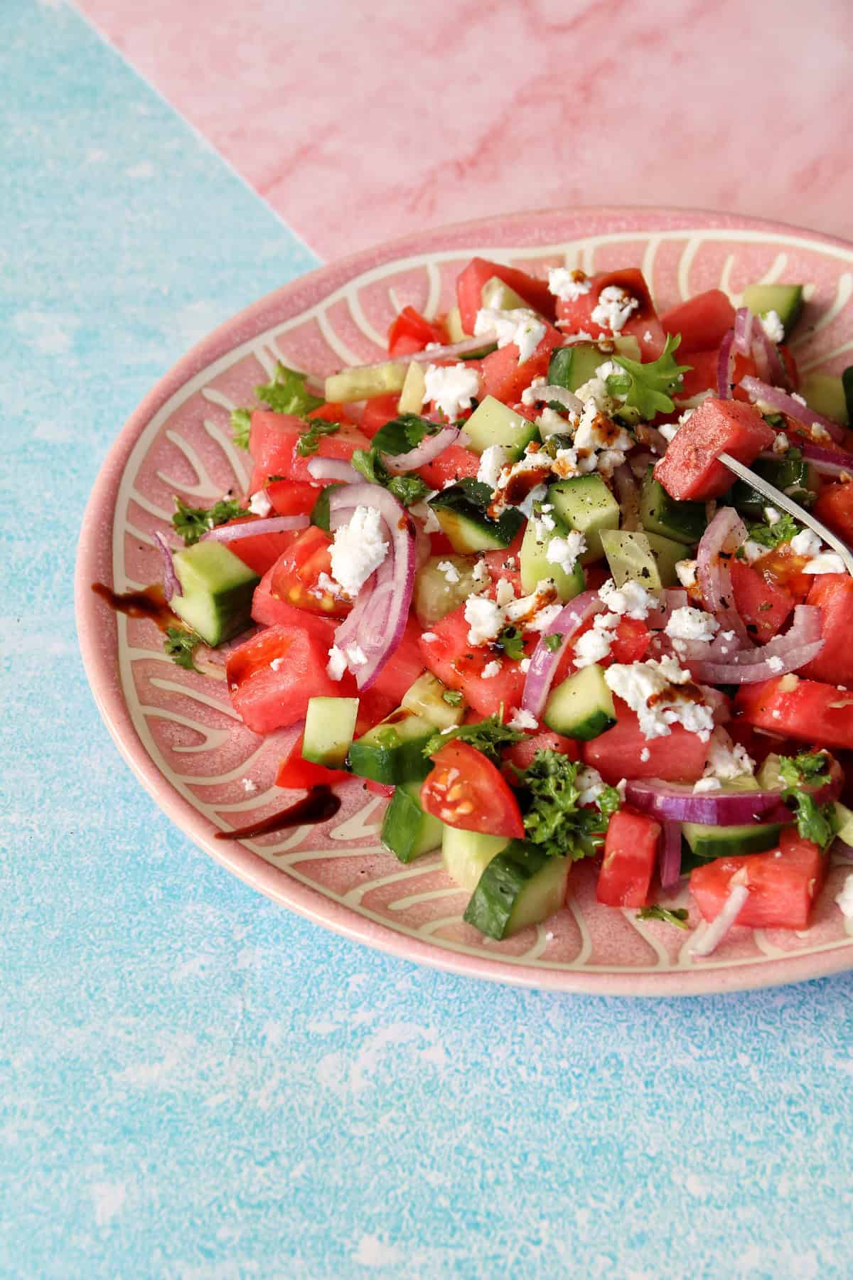 Detailaufnahme der Zutaten vom Salat, deutlich zu erkennen: rote Zwiebeln, Gurken, Tomaten, Wassermelone und Feta. Mit Balsamico beträufelt.