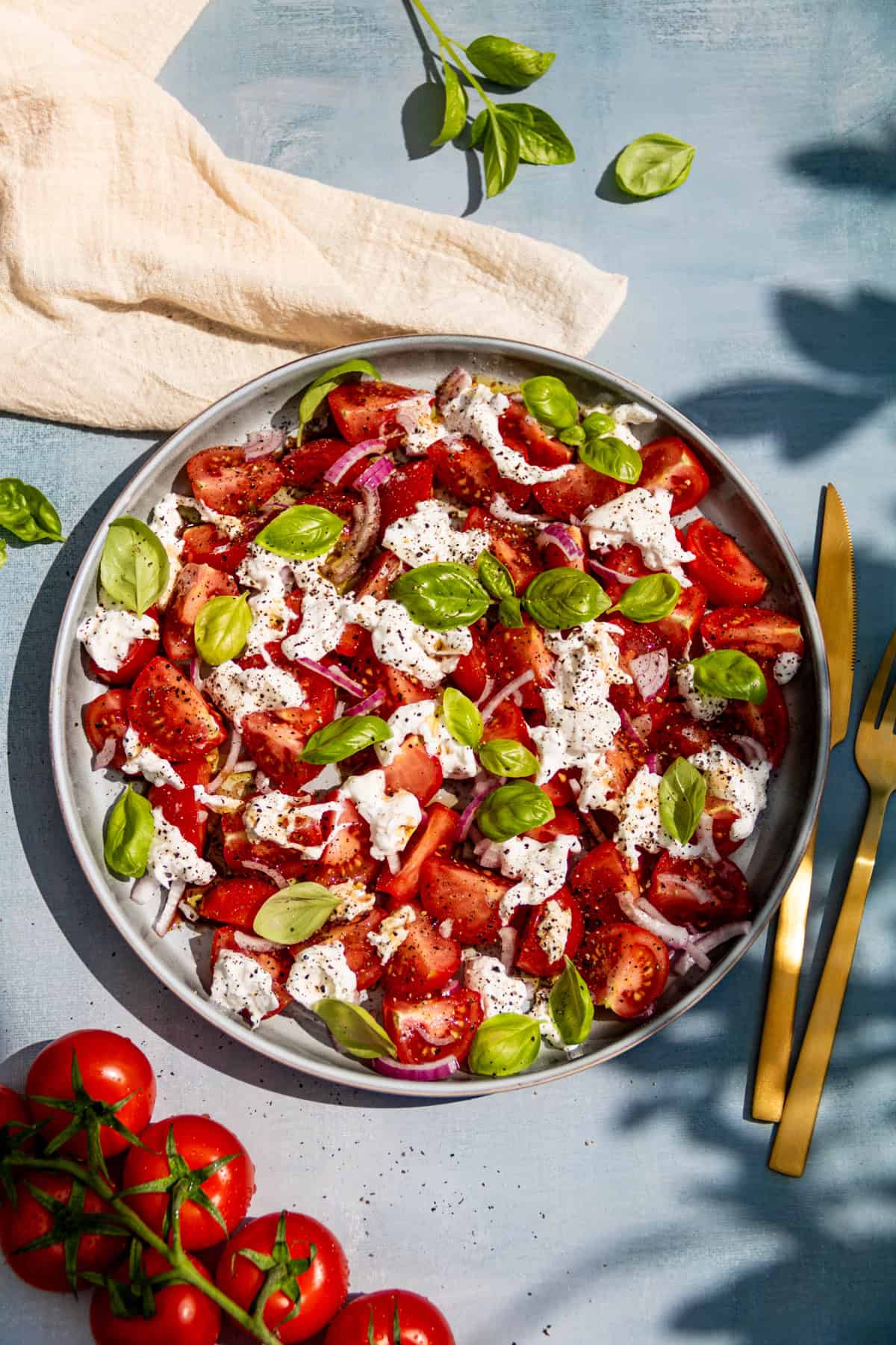 Salatteller von oben fotografiert. Sommerlich präsentiert. Goldenes Besteck liegt neben dem Tomaten-Burrata-Salat.