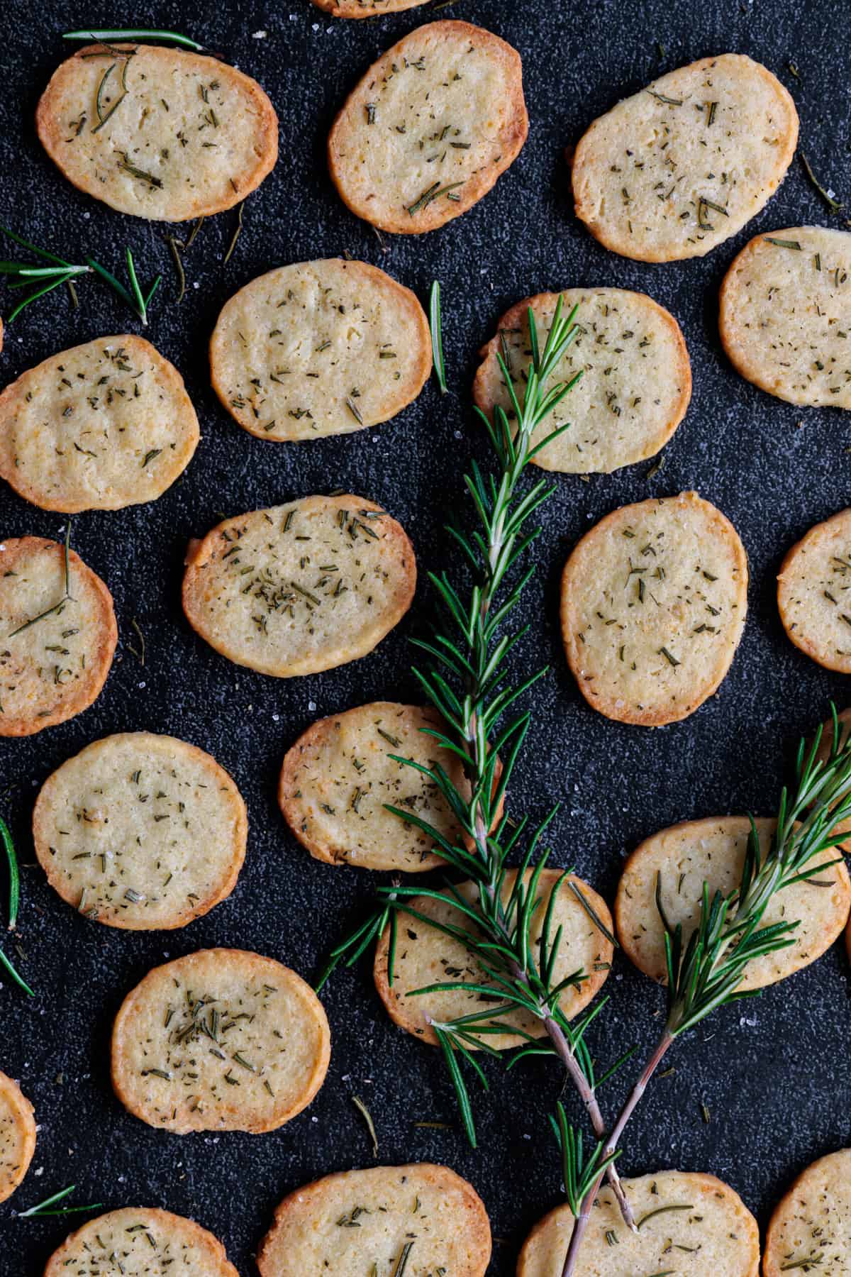 Der perfekte salzige Snack: Parmesan-Rosmarin-Cracker liegen auf einem dunklen Untergrund willkürlich nebeneinander. Darauf liegen 2 Zweige frischer Rosmarin.