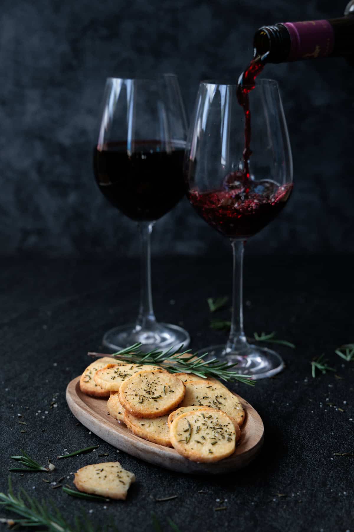 Ein paar salzige Parmesan-Rosmarin-Cracker werden auf einem Tablett mit einem Glas Wein serviert.