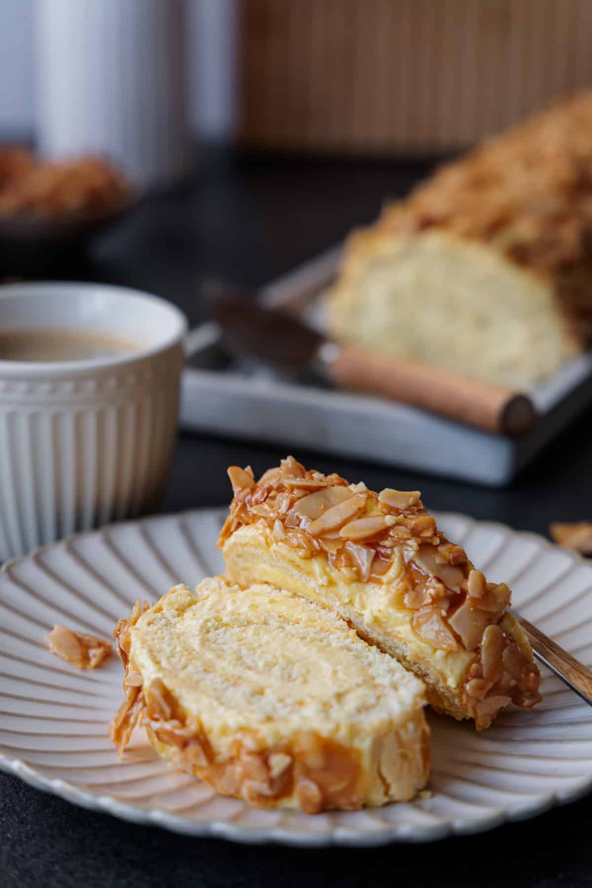 2 Stücke der Biskuitroulade auf einem wellenförmigen Kuchenteller. Der Honig-Mandel Belag à la Bienenstich-Kuchen ist sehr appetitlich in Szene gesetzt.