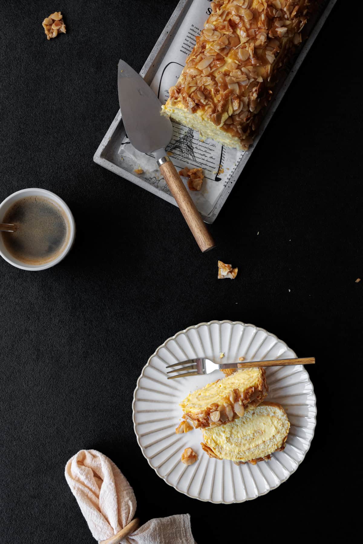 Die Biskuitrolle auf der Tortenplatte ist oben zu sehen, weiter unten ein Teller mit dem Bienenstich-Kuchenstück. Eine Tasse Kaffee steht ebenfalls im Bild.