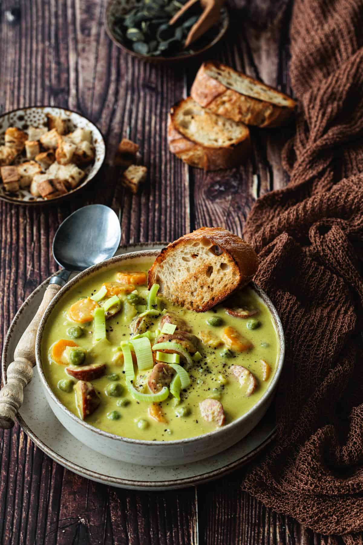 Erbseneintopf wie bei Oma in einer tiefen Schale. Mit angerösteten Baguettescheiben und Croûtons. Ein paar Porrestücke und frischer Pfeffer dekorieren die Erbsensuppe.