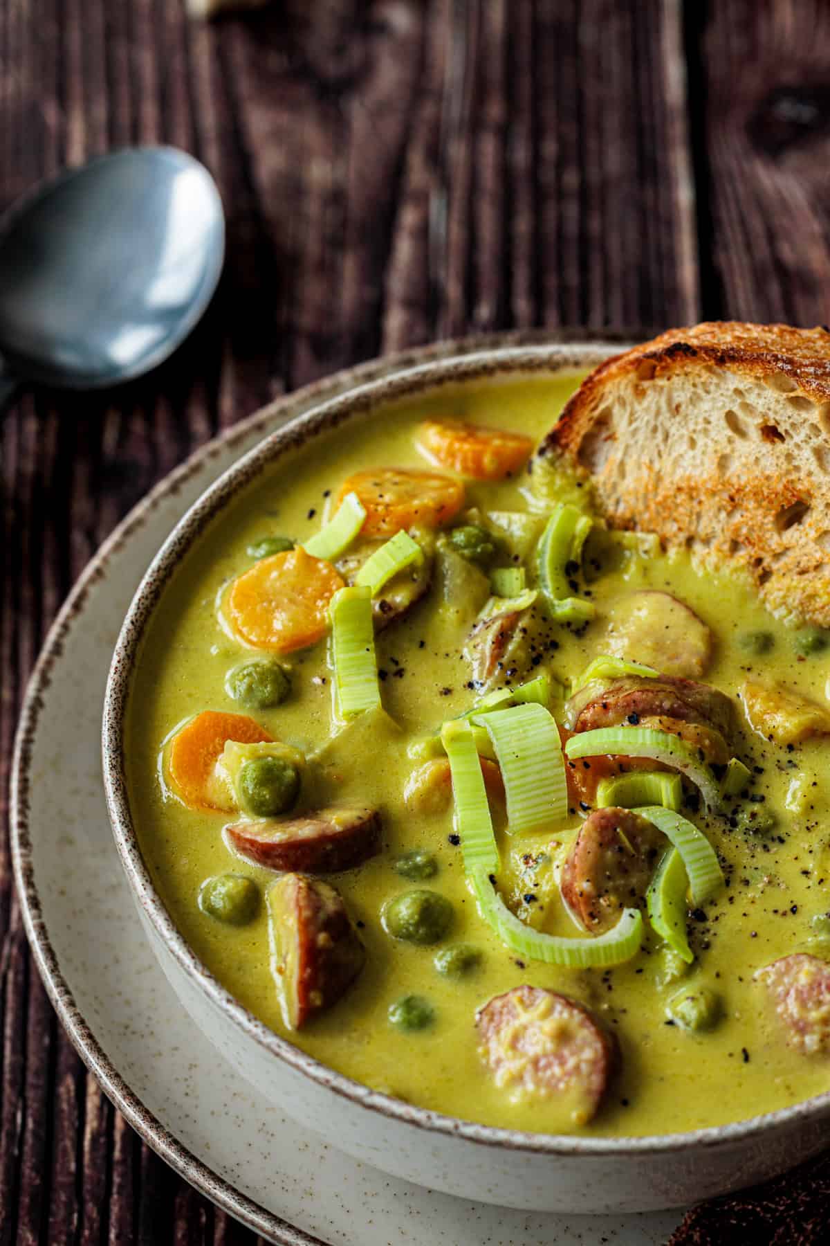 Nahaufnahme des Erbseneintopfes. Mettenden, Möhren, Kartoffeln und Erbsen sind gut zu erkennen. Eine angeröstete Baguettescheibe ist in die Erbsensuppe getaucht.