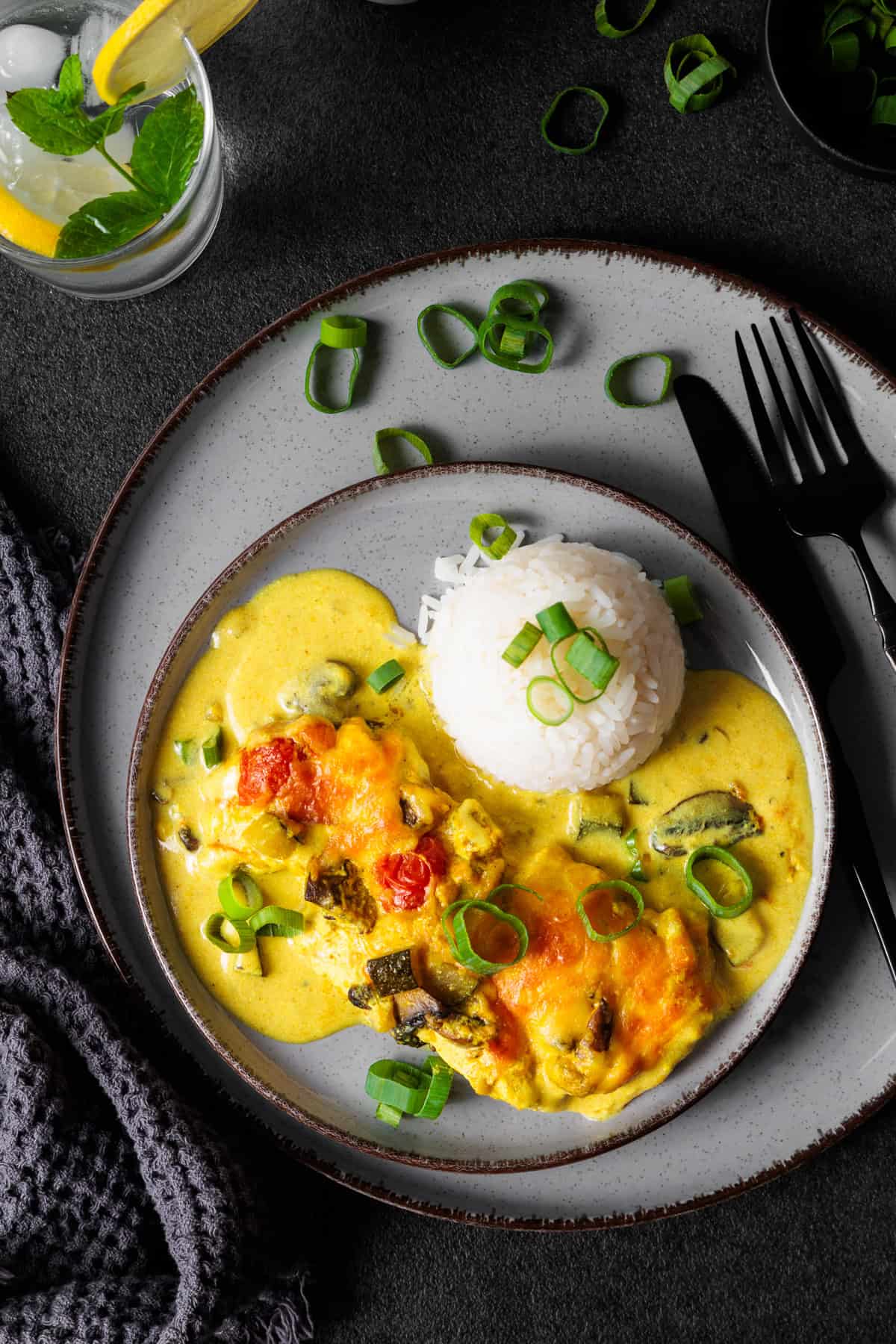 Schnitzel-Gratin mit Curry und Gemüse auf einem grauen Teller. Daneben liegt schwarzes Besteck und im Hintergrund Frühlingszwiebeln und ein Glas mit Zitronenwasser.