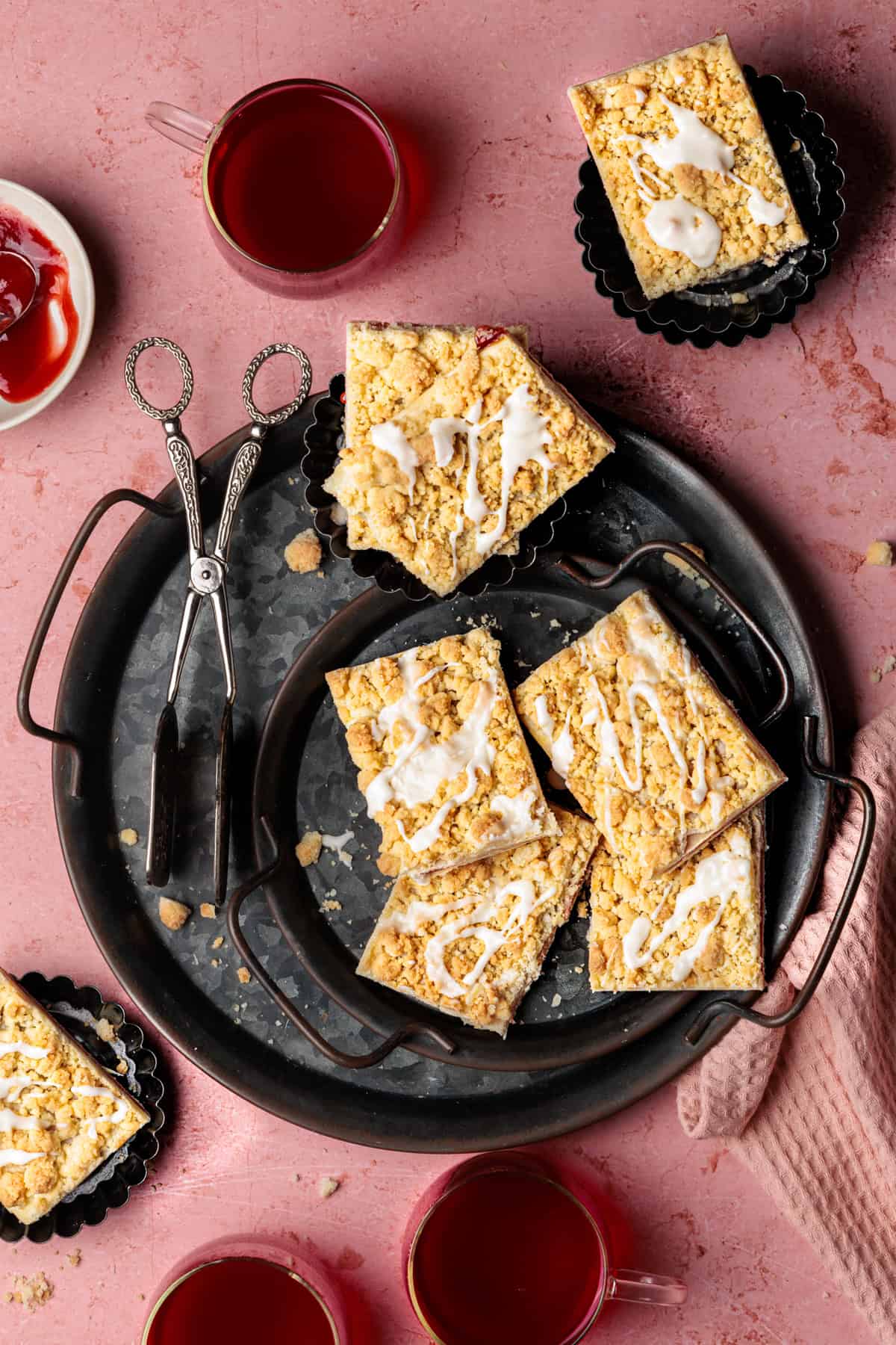 Auf einem rosafarbenen Hintergrund liegen Stücke eines goldbraun gebackenen Prasselkuchens aus Blätterteig. Sie sind mit Streuseln und einem Hauch von Glasur verziert. Die Füllung ist eine Erdbeermarmelade. Die Kuchenstücke liegen auf einem Metallteller.