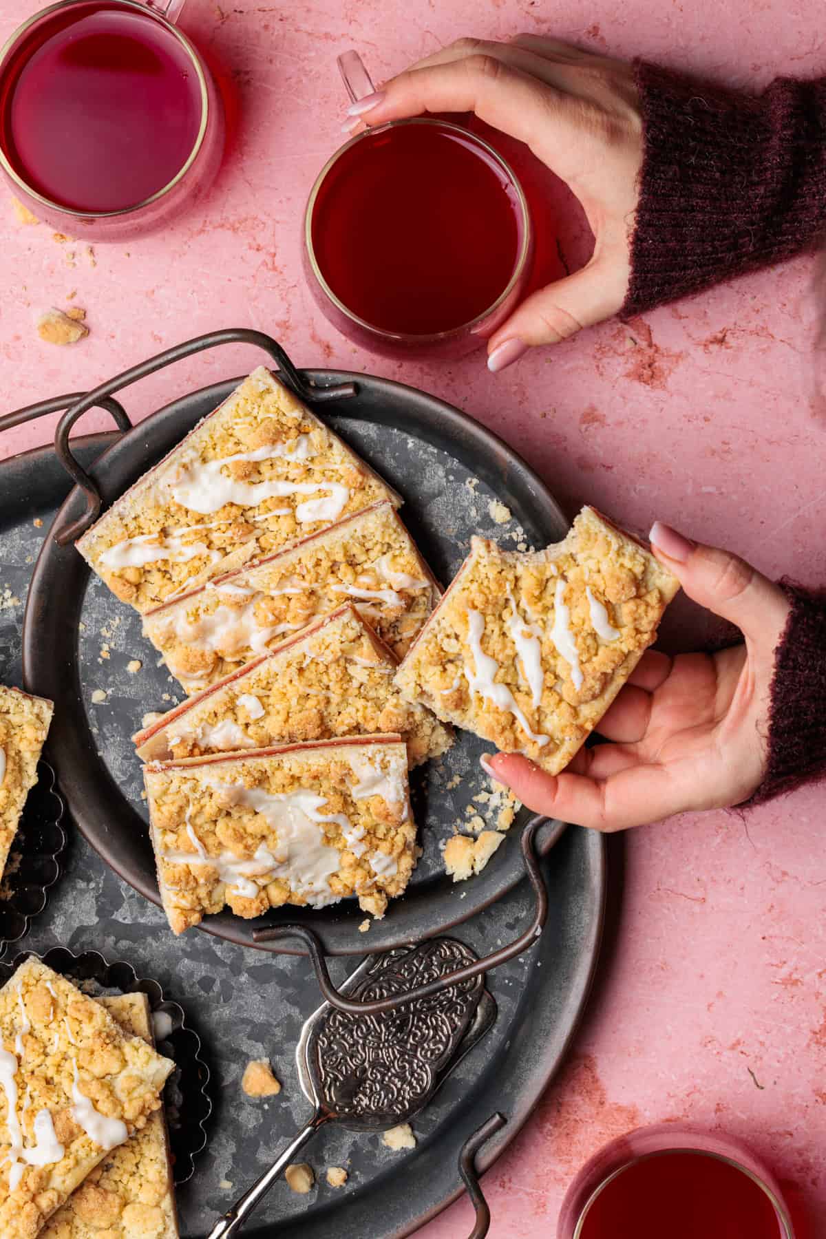 Warme Hände umschließen ein Stück goldbraunen Prasselkuchens aus Blätterteig mit Marmeladenfüllung und Streuseln, während dampfender Tee in Gläsern daneben steht. Das Foto strahlt eine gemütliche Atmosphäre aus und lädt dazu ein, sich eine kleine Auszeit zu gönnen.