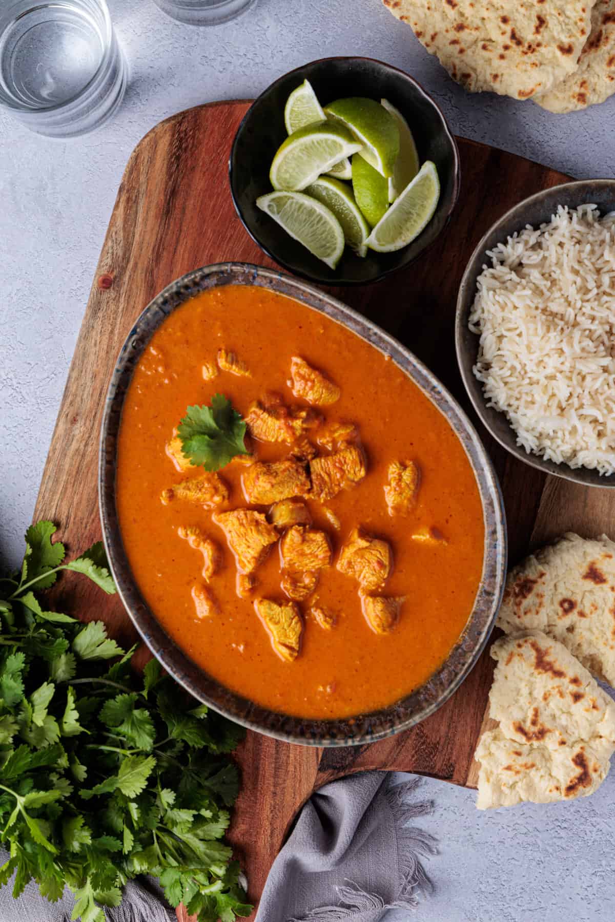 Rezept für Butter Chicken mit Tikka Masala Paste aus dem Thermomix® - die Soße befindet sich in einer ovalen Schale. Diese steht auf einem Holzbrett. Daneben liegt ein Bund Koriander, eine Schale Reis, ein Schälchen mit Limettenstücken und indischem Naanbrot.