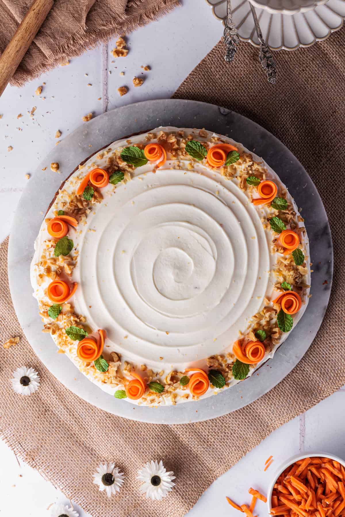 Saftiger Karottenkuchen mit Frischkäse-Topping (Thermomix®-Rezept) von oben fotografiert. Den Rand der Torte schmücken Rosen aus aufgerollten Möhrenstreifen sowie gehackte Walnüsse und Minzblättchen. Der Möhrenkuchen steht auf einer Marmorplatte. Daneben sind braune Tischdecken/Servietten zu sehen.