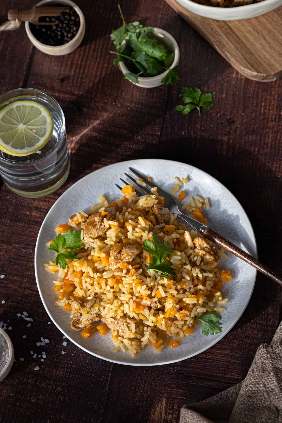 Ein Tischarrangement mit dem Hauptmotiv Plov auf die Schnelle aus dem Thermomix® auf dem Teller, mit frischer Petersilie garniert. Eine Gabel mit Holzgriff liegt auf dem Teller. Daneben ein Glas Wasser mit Zitrone.