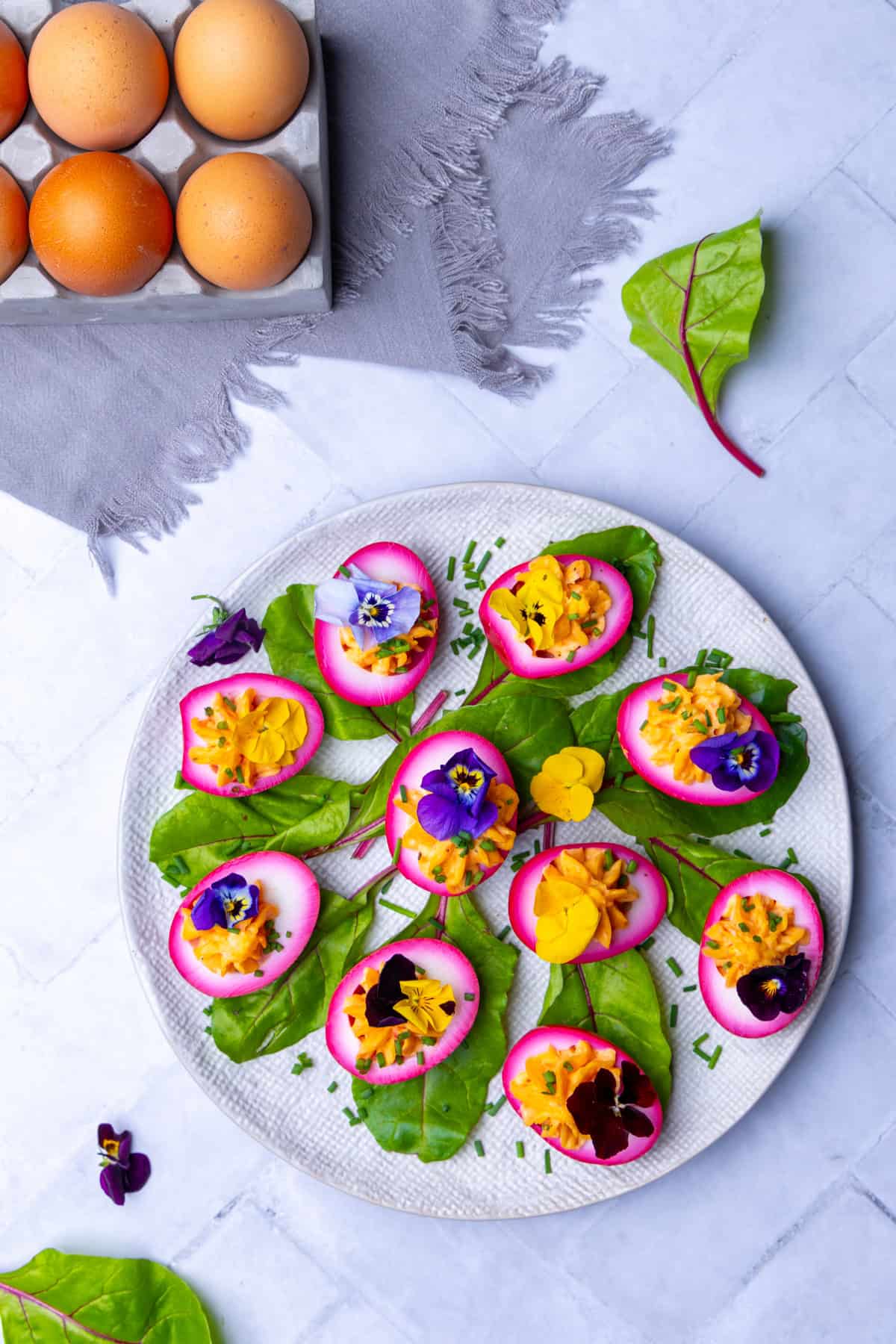 Osterbrunch - Idee mit gekochten Eiern: Ein Teller mit bunten, gefüllten Rote Beete Eiern, verziert mit essbaren Blüten und Kräutern, daneben ein Eierkarton mit ungekochten Eiern und eine graue Stoffserviette. Rezept auch für den Thermomix® geeignet.