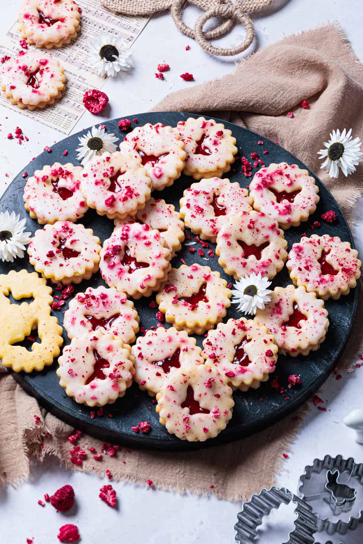 Eine rustikale schwarze Marmorplatte, gefüllt mit frisch gebackenen Himbeer-Linzer-Keksen. Die goldbraunen Osterkekse sind mit einer zarten weißen Puderzucker-Glasur überzogen und mit roten getrockneten Himbeerstückchen dekoriert. Zwischen den Keksen liegen weiße Blüten und ein Ausstecher in Form eines Hasen.