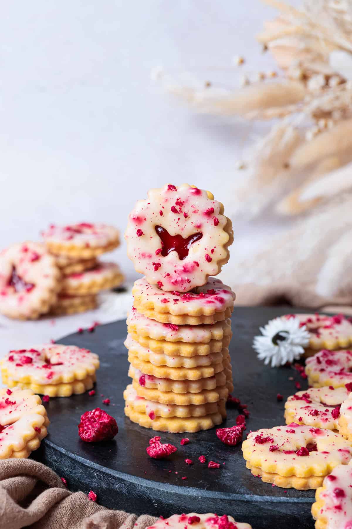 Ein Stapel selbstgebackener Himbeer-Linzer-Kekse auf einem dunklen Steinteller. Die goldbraunen Kekse sind mit einer zarten weißen Glasur überzogen und mit roten Himbeerstückchen dekoriert. Im Hintergrund sind getrocknete Blumen zu sehen.