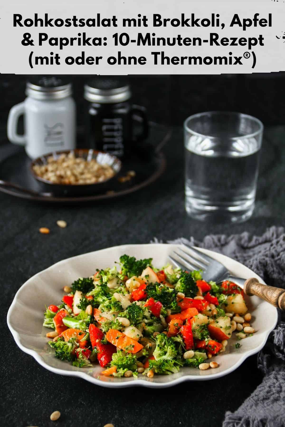 Pin zu Pinterest: Draufsicht auf einen angerichteten Rohkostsalat (mit oder ohne Thermomix® zubereitet) auf einem runden, hellen Teller, der auf einer dunklen Arbeitsfläche steht. Der Salat besteht aus fein gehacktem Brokkoli, roten Paprikawürfeln, Apfelstückchen und gerösteten Pinienkernen. Eine Gabel liegt neben dem Teller. Im Hintergrund stehen zwei kleine Gläser mit der Aufschrift 'SALT' und 'PEPPER', eine kleine Schale mit Pinienkernen und ein Glas mit Wasser. Ein graues Tuch ist locker neben dem Teller drapiert.