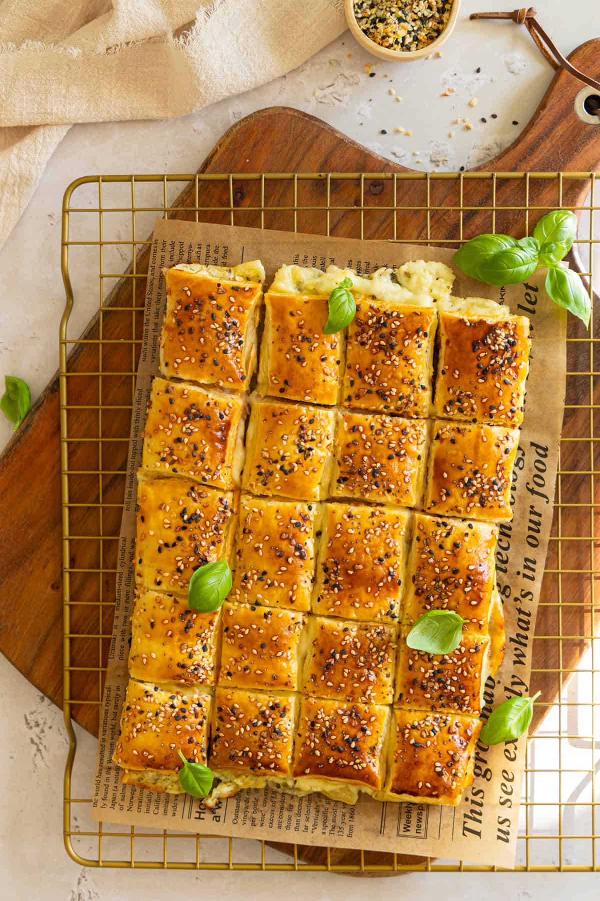  Nahaufnahme eines goldbraunen Käse-Schinken-Zupfbrots aus Blätterteig, serviert auf einem goldenen Drahtgestell mit Backpapierunterlage und Holztisch im Hintergrund. Das Zupfbrot ist in quadratische Portionen geschnitten, mit Sesam bestreut und mit frischen Basilikumblättern garniert. Im oberen Bereich ist eine kleine Schale mit Sesammischung zu sehen. Perfekt für schnelle herzhafte Blätterteig Snacks und einfache Partyrezepte.