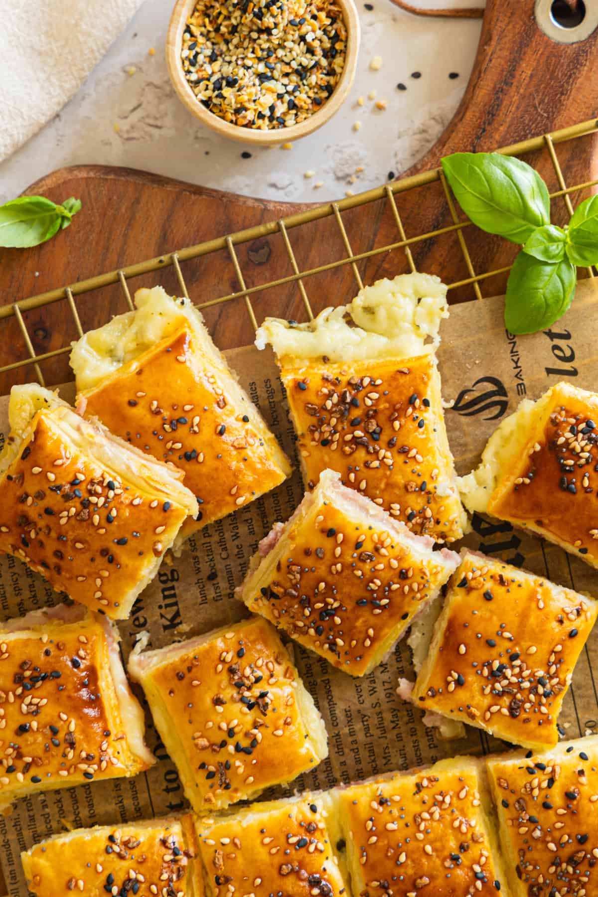 etailaufnahme von einzelnen Stücken eines goldbraunen Käse-Schinken-Zupfbrots aus Blätterteig, die auf einem goldenen Drahtgestell mit Backpapierunterlage und einem Holztisch im Hintergrund liegen. Die quadratischen Stücke sind mit Sesam bestreut und zeigen die Füllung aus Käse und Schinken. Frische Basilikumblätter dienen als Garnitur. Im oberen Bereich ist eine kleine Schale mit Sesammischung zu sehen. Ideal für einfache herzhafte Blätterteig Snacks für Partys und schnelle Rezepte für Gäste.