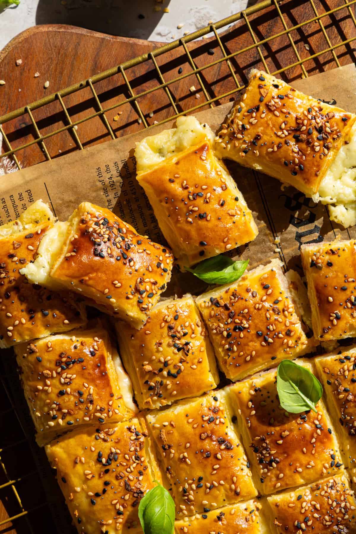 Draufsicht auf ein Käse-Schinken-Zupfbrot aus Blätterteig, das auf einem goldenen Drahtgestell mit Backpapierunterlage und einem Holztisch im Hintergrund präsentiert wird. Die einzelnen Quadrate des Zupfbrots sind deutlich erkennbar, goldbraun gebacken, mit Sesam bestreut und mit frischen Basilikumblättern garniert. Das Bild zeigt die Textur des knusprigen Blätterteigs und die Füllung aus geschmolzenem Käse und Schinken. Ideal für schnelle einfache Partyrezepte und herzhafte Blätterteig Snacks für Geburtstage.