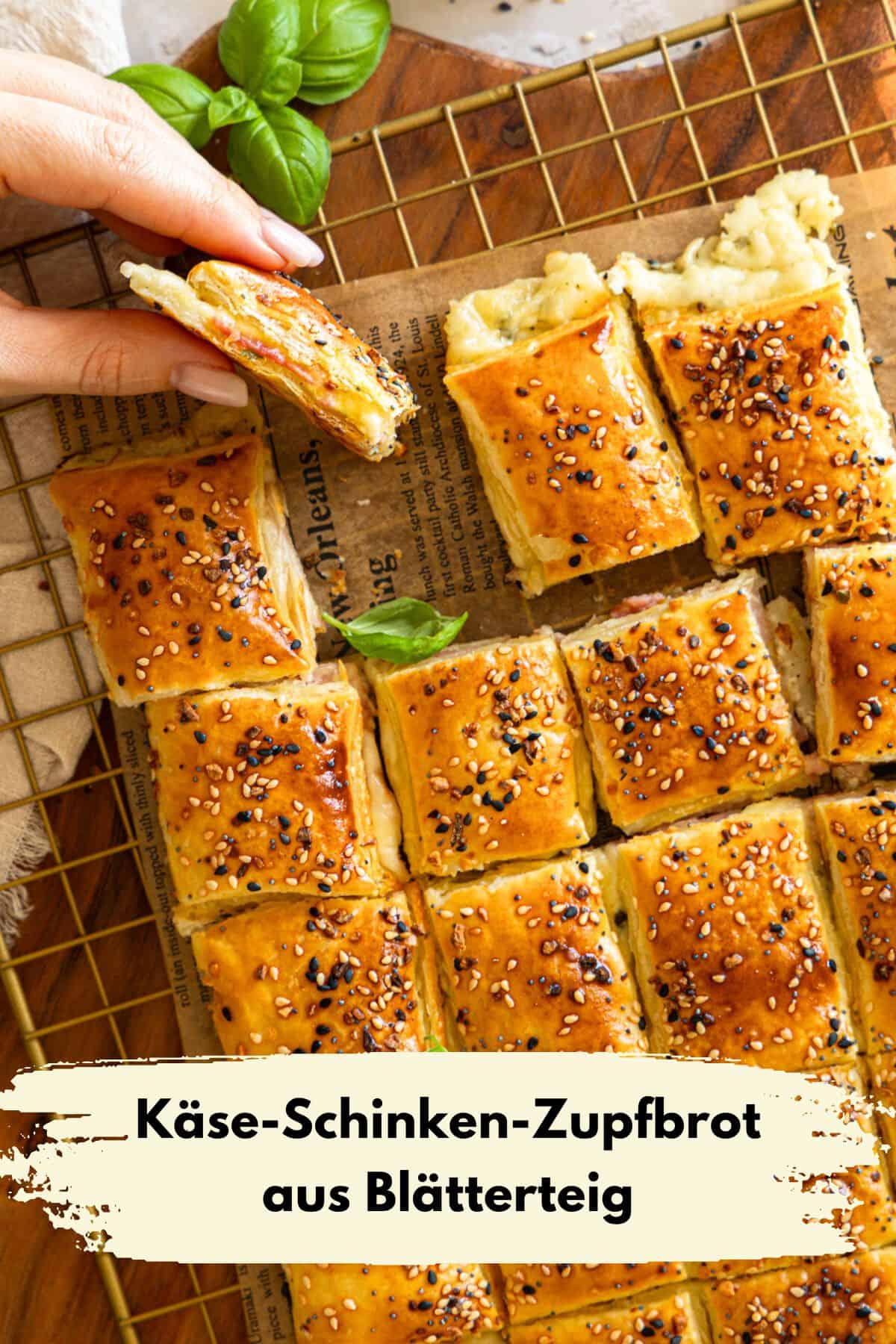 Pin zu Pinterest: Hand, die ein Stück goldbraunes Käse-Schinken-Zupfbrot aus Blätterteig vom goldenen Drahtgestell hebt. Das Zupfbrot ist in Quadrate geschnitten, mit Sesam bestreut und zeigt die Füllung aus Käse und Schinken. Im Hintergrund liegt das restliche Zupfbrot auf Backpapier mit Zeitungsmuster und ist mit frischen Basilikumblättern garniert. Ideal für einfache herzhafte Blätterteig Snacks für Geburtstage und schnelle Rezepte für Gäste.