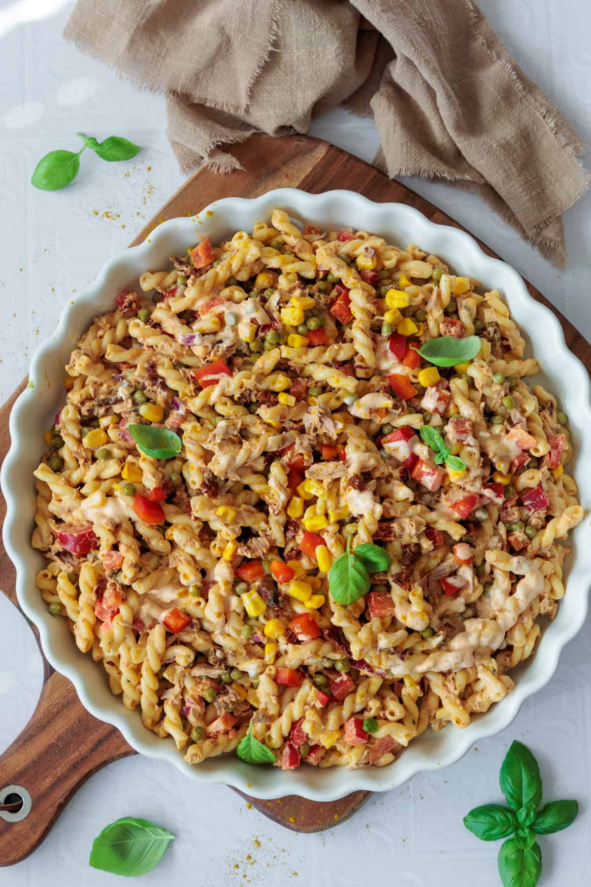 Draufsicht auf einen bunten Thunfischsalat mit Nudeln in einer weißen, runden Auflaufform auf einem Holzbrett. Der Salat enthält Spiralnudeln, Thunfischstücke, gelben Mais, rote Paprikawürfel und grüne Erbsen. Frische Basilikumblätter sind als Dekoration auf dem Salat und dem Brett verteilt. Ein beigefarbenes Tuch liegt am oberen Bildrand. 