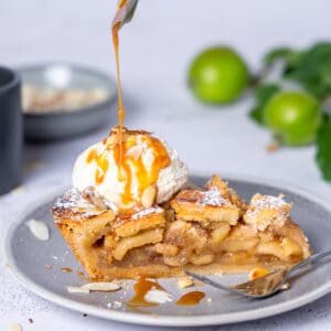 Nahaufnahme eines angeschnittenen Stücks American Apple Pie (Apfelkuchen) auf einem grauen Teller. Das Kuchenstück zeigt die mürbe Kruste und die saftige Apfelfüllung. Es ist garniert mit einer Kugel Vanilleeis, Mandelsplittern und einem Drizzle Karamellsauce, die gerade von einem Löffel gegossen wird.