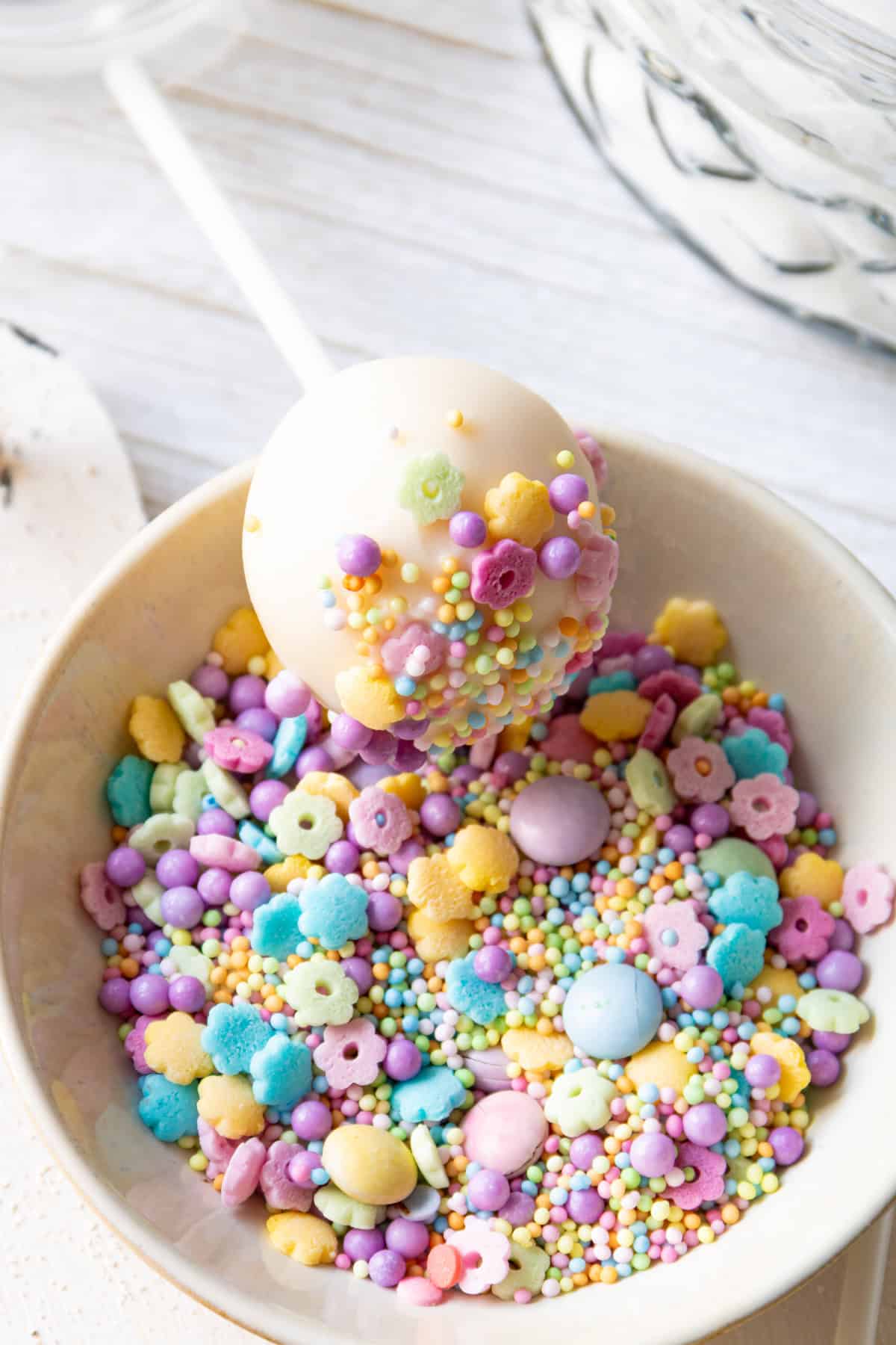 Ein weiß glasierter Cake Pop mit einem Stiel wird über einer Schüssel voller pastellfarbener Streusel und bunter Zuckerperlen gehalten. Er zeigt die Dekoration, die für das einfache Cake-Pop-Rezept aus dem Thermomix® für Kindergeburtstage verwendet wird.