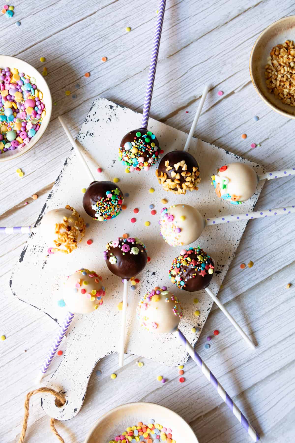 Das Bild zeigt eine Draufsicht auf neun appetitliche Cake Pops, die auf einem rustikalen weißen Holzbrett ausgelegt sind. Sie sind mit Kuchenglasur in Zartbitter und Vanille überzogen, was eine schöne Farbvielfalt erzeugt. Einige der Kuchenkugeln am Stiel sind mit gehackten Nüssen oder Krokant dekoriert, während andere mit einer bunten Mischung aus Streuseln und bunten Zuckerperlen verziert sind. Einige der Cake-Pop-Stiele sind weiß, andere lila-weiß gestreift, was dem Bild eine spielerische Note verleiht. Im oberen linken Bereich des Bildes ist eine kleine Schüssel mit weiteren farbenfrohen Streuseln zu sehen, während oben rechts eine Schüssel mit gehackten Nüssen oder Krokant platziert ist, bereit für die Dekoration. Einzelne verstreute Streusel liegen auf dem hellen Holzhintergrund und verstärken den Eindruck eines kreativen Backprozesses für diese einfachen und beliebten Leckereien, die perfekt für kleine Kinderhände sind und leicht mit dem Thermomix® zubereitet werden können.
