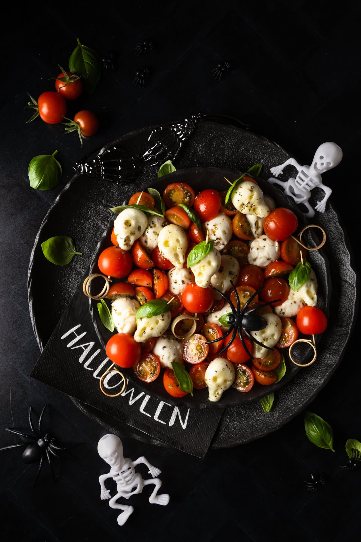 Du siehst auf dem Bild einen Teller mit einem Halloween-Caprese-Salat. Der Teller ist dunkel, vermutlich schwarz oder anthrazit, und hat einen erhöhten, leicht unebenen Rand, was ihm eine rustikale Optik verleiht.

Im Mittelpunkt des Bildes befindet sich der Salat. Er besteht hauptsächlich aus halbierten Kirschtomaten in verschiedenen Rottönen und gelblichen Mozzarella-Totenköpfen, die in Silikonformen gegossen wurden. Zwischen den Tomaten und den Mozzarella-Kugeln sind frische, grüne Basilikumblätter verteilt.

Einige der Zutaten sind zu Spießen arrangiert, die in den Salat gesteckt sind. Die Mozzarella-Totenköpfe sind dabei fest auf den Spießen, abwechselnd mit Tomaten und Basilikum.

Um den Teller herum sind verschiedene Halloween-Deko-Elemente drapiert. Auf der linken Seite liegen eine schwarze Plastikspinne und ein kleines weißes Skelett. Auf der rechten Seite sind ebenfalls ein kleines Skelett und eine weitere schwarze Spinne zu sehen.