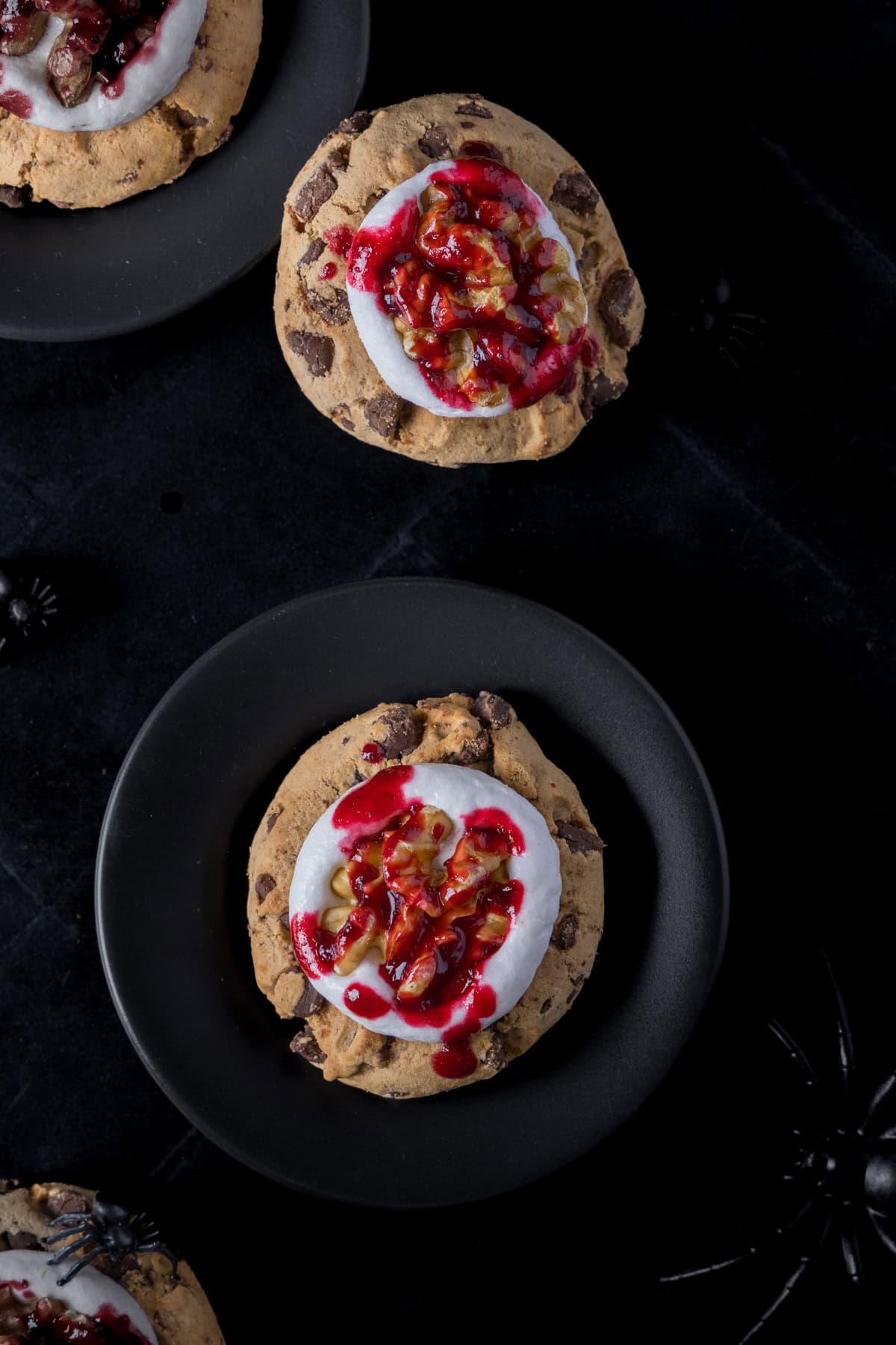 Blutige Gehirnkekse für Halloween auf einem schwarzen kleinen Teller. Die Schichten -bestehend aus Cookies, Marshmallow und Walnusshälfte mit roter Marmelade sind deutlich zu erkennen. Schwarze Plastikspinnen laufen durchs Bild.