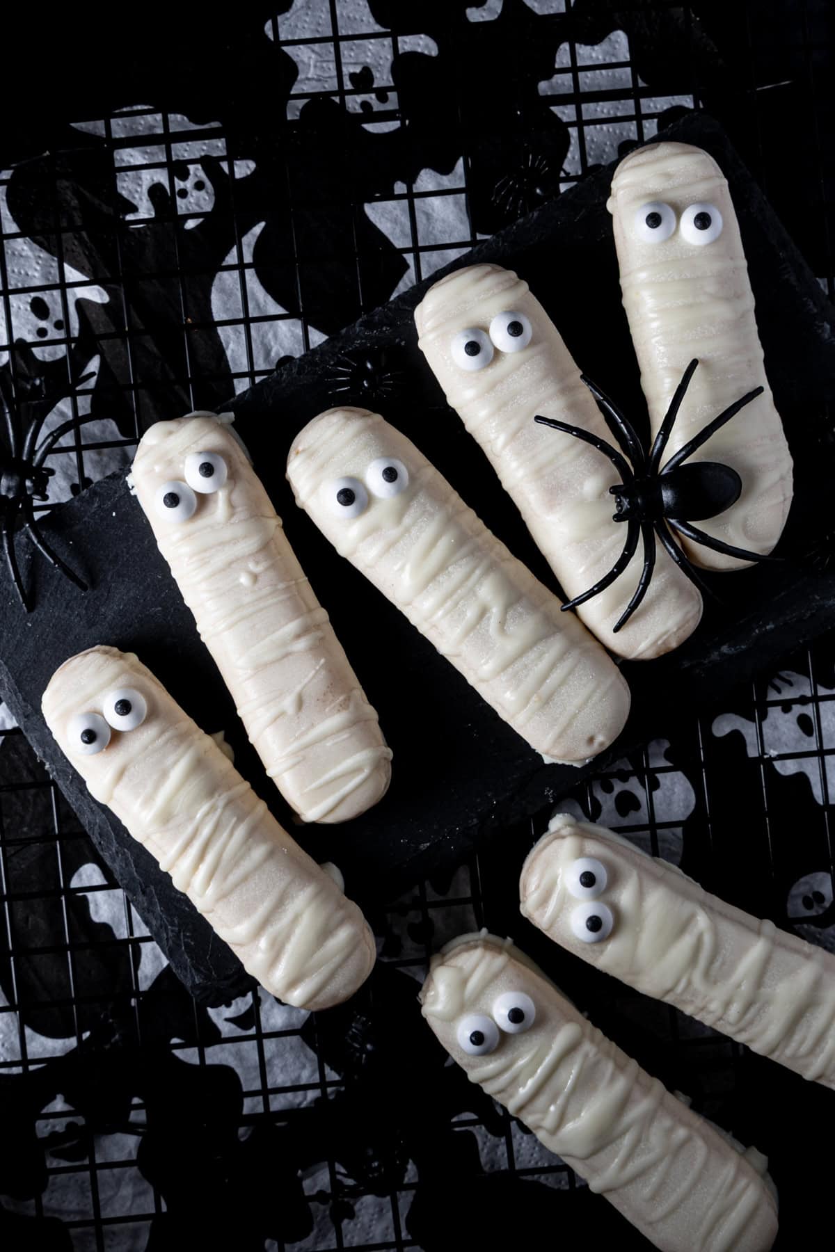 Fingerfood für die Kinderhalloween-Party: Löffelbiskuit-Mumien mit weißer Schokolade überzogen, auf einem schwarzen Hintergrund mit Gespensterserviette und Plastikspinne.