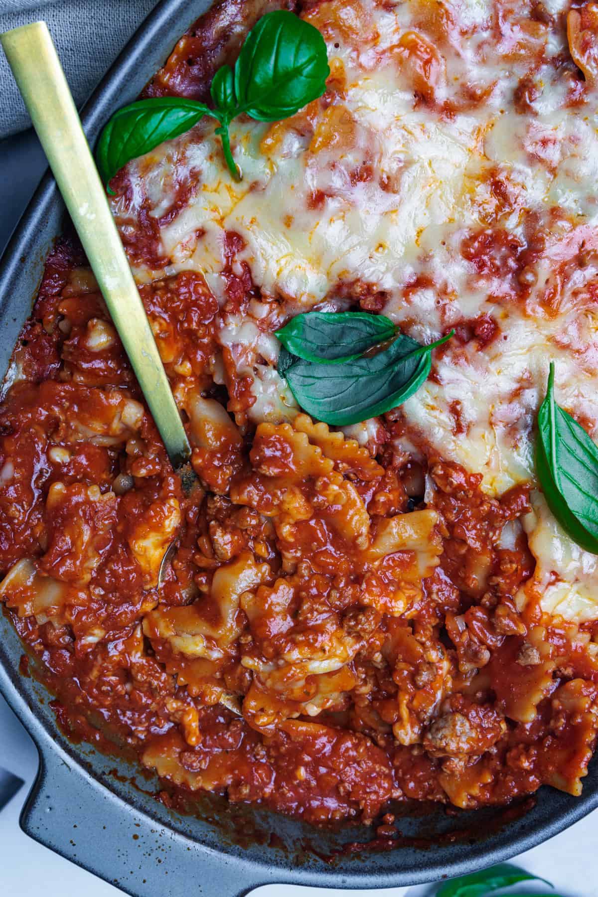 Ein detaillierter Nahaufnahme-Schuss eines Nudelauflaufs mit Hackfleisch, der in einer ovalen, grauen Auflaufform serviert wird. Ein goldener Löffel steckt im Gericht. Man sieht die Farfalle-Nudeln in der herzhaften Tomaten-Hackfleisch-Soße. Die obere Hälfte des Auflaufs ist mit einer Schicht aus geschmolzenem, goldbraunem Käse bedeckt und mit frischen Basilikumblättern garniert.