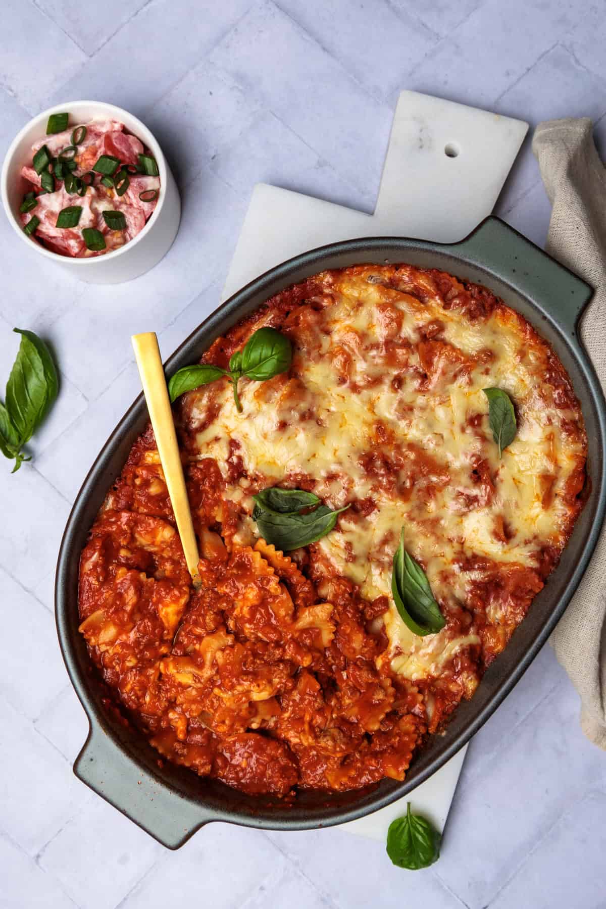 Ein köstlicher Auflauf aus Schleifchennudeln, Hackfleisch, Tomatensauce und geschmolzenem Käse, verfeinert mit Basilikum. Er wird in einer ovalen Auflaufform serviert, daneben steht eine kleine Schüssel mit Tomatensalat. Das Bild zeigt eine Nahaufnahme des herzhaften Ofengerichts, das als schnelles und familienfreundliches Rezept beschrieben wird.