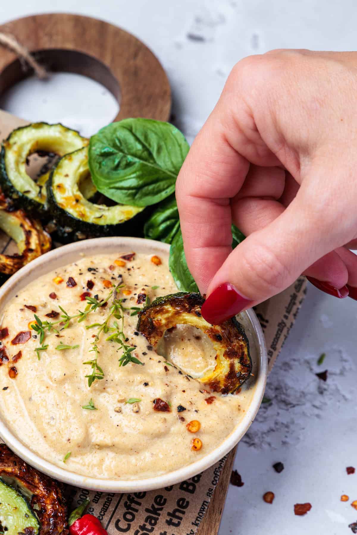 Nahaufnahme einer weiblichen Hand mit roten Fingernägeln, die eine gebratene Zucchinischeibe in einen cremigen, mit Chiliflocken und frischem Thymian bestreuten Dip taucht. Im Hintergrund sind weitere Zucchinischeiben und ein Stück Holzschneidebrett zu sehen.