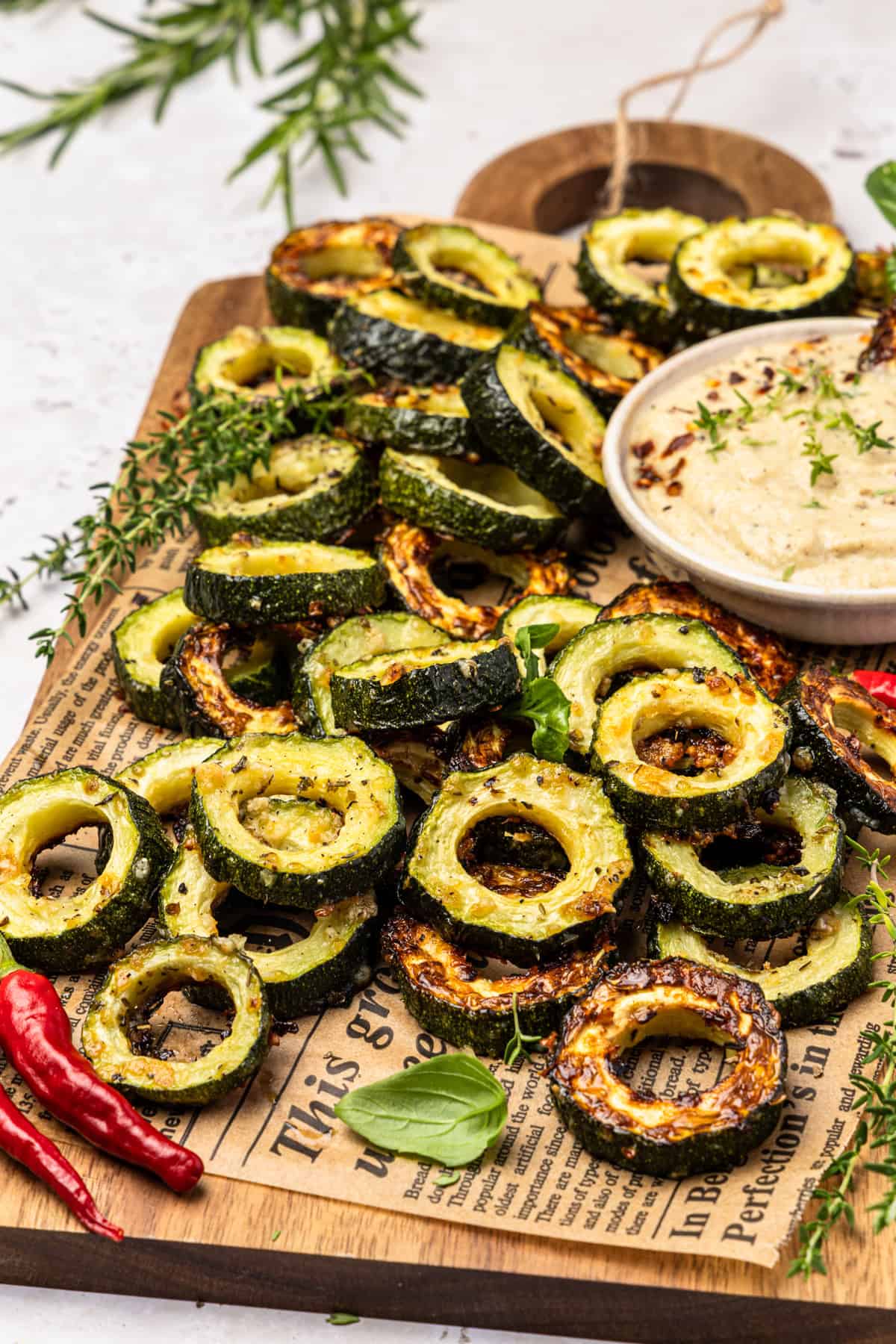 Gebackene, in Scheiben geschnittene Zucchini-Scheiben mit Parmesan und italienischen Kräutern, angerichtet auf einem Schneidebrett mit Zeitungspapier. Dazu wird ein cremiger Dip aus den Zucchini-Mitten, Knoblauch, Schmand, Zitronensaft und Gewürzen in einer Schale serviert. Das Gericht ist mit frischen Kräutern garniert.