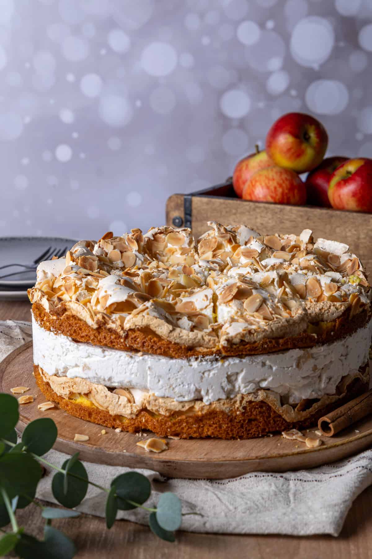 Bratapfel-Torte mit knuspriger Baiser-Haube und Mandeln. Die zweischichtige Torte zeigt eine cremige Füllung und eine sichtbare Bratapfel-Kompott-Schicht zwischen den Böden. Im Hintergrund liegen frische Äpfel in einer Holzkiste und Zimtstangen auf einem rustikalen Holzbrett, vor einem weichgezeichneten, festlichen Lichterglanz. Perfektes Thermomix®-Rezept für Weihnachten und die kalte Jahreszeit.