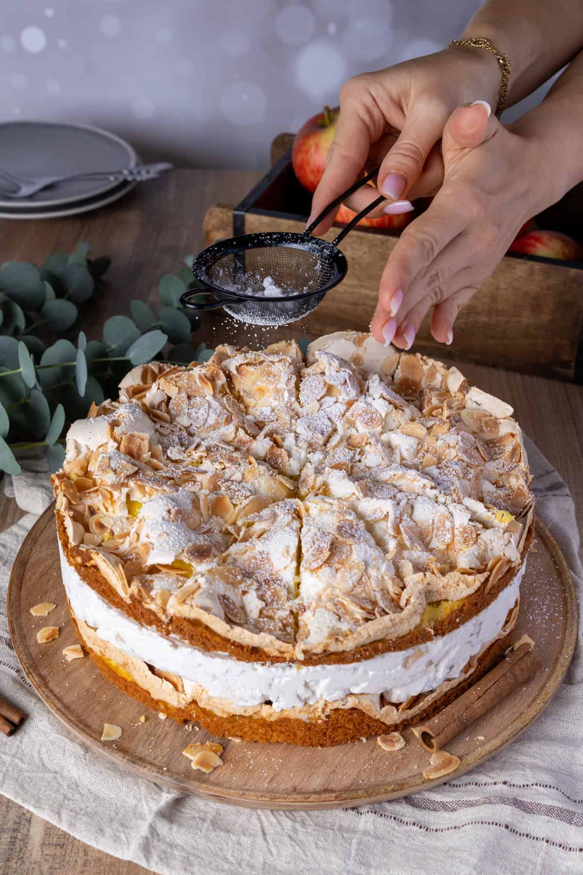 Hände pudern eine fertig geschnittene Bratapfel-Torte mit Baiser-Haube und Mandeln mit Puderzucker. Die cremige Füllung und die Bratapfel-Kompott-Schicht sind durch die Schnitte sichtbar. Im Hintergrund sind frische Äpfel und Zimtstangen auf einem rustikalen Holzbrett zu sehen. Ein festliches Thermomix®-Rezept für die kalte Jahreszeit.