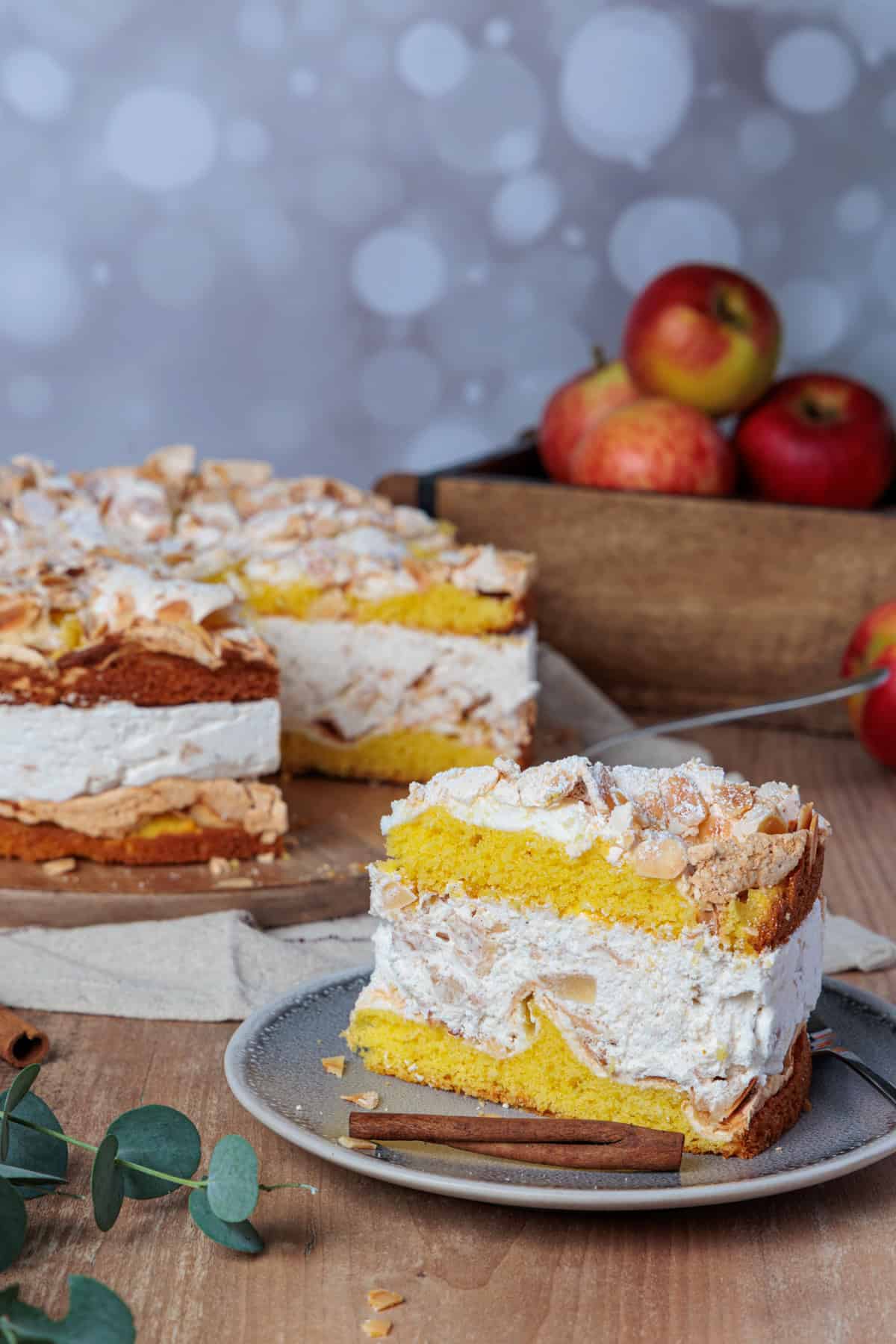 Ein großes Stück Bratapfel-Torte mit Baiser-Haube und Mandeln auf einem Teller, daneben die angeschnittene Torte im Hintergrund. Die einzelnen Schichten sind gut sichtbar: goldener Rührteig, eine dicke Schicht cremiger Füllung und die Bratapfel-Kompott-Schicht mit Apfelstücken. Im Hintergrund liegen frische Äpfel in einer Holzkiste und Zimtstangen auf einem rustikalen Holzbrett, vor einem weichgezeichneten Lichterglanz. Ideal als festliches Dessert zur Weihnachtszeit, zubereitet im Thermomix®.