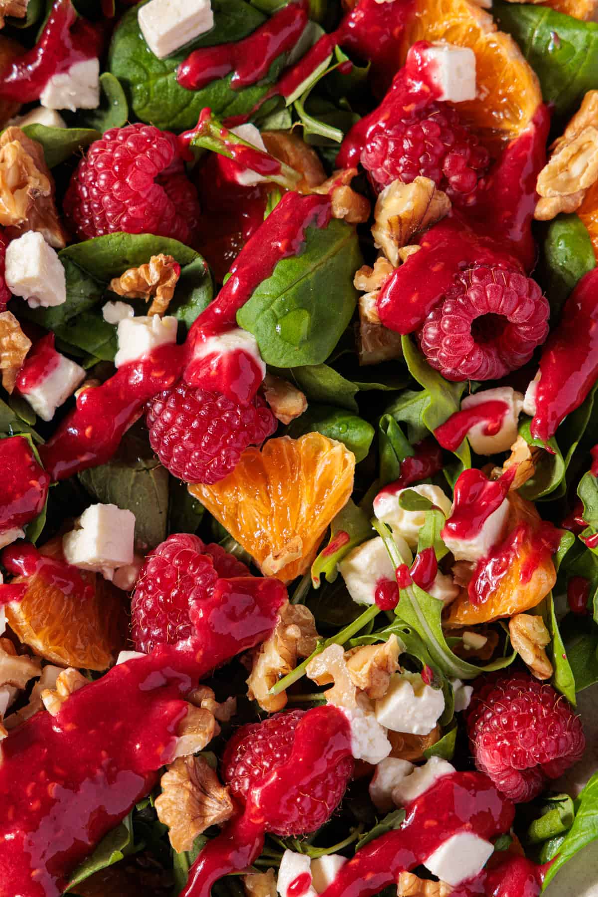 Ein Close-up-Foto zeigt einen bunten Feldsalat, der mit verschiedenen Zutaten und einem Dressing vermischt ist. Man sieht Orangenfilets, ganze Himbeeren, gewürfelten Feta-Käse und gehackte Walnüsse. Ein leuchtend rotes Himbeerdressing ist großzügig über die Zutaten geträufelt. Die glänzenden Blätter des Feldsalats und des Rucola sind die Basis des Gerichts. Das Bild ist aus der Vogelperspektive aufgenommen und füllt den gesamten Rahmen.